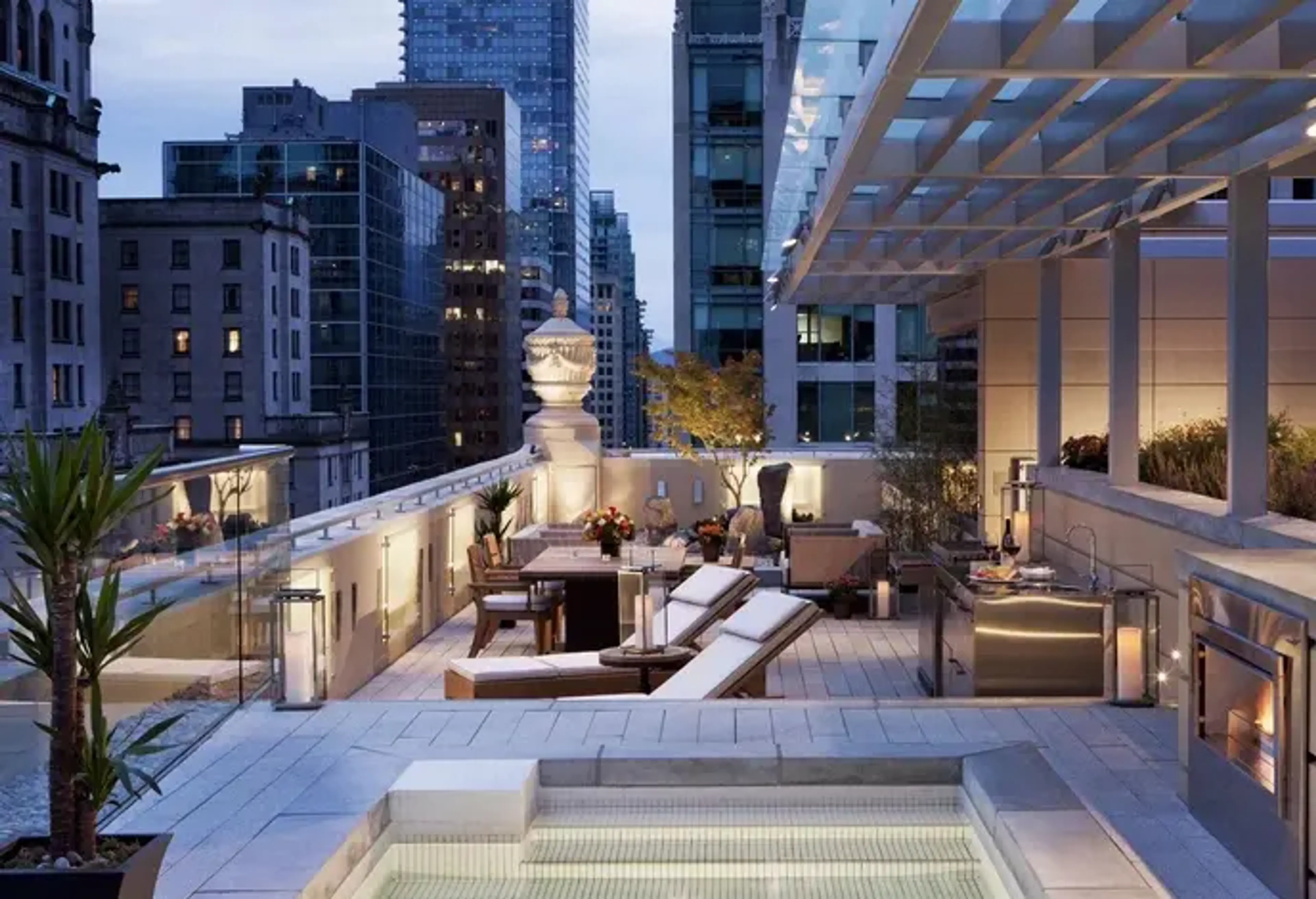 Modern rooftop terrace with lounge chairs, dining table, plants, and city skyline at dusk.