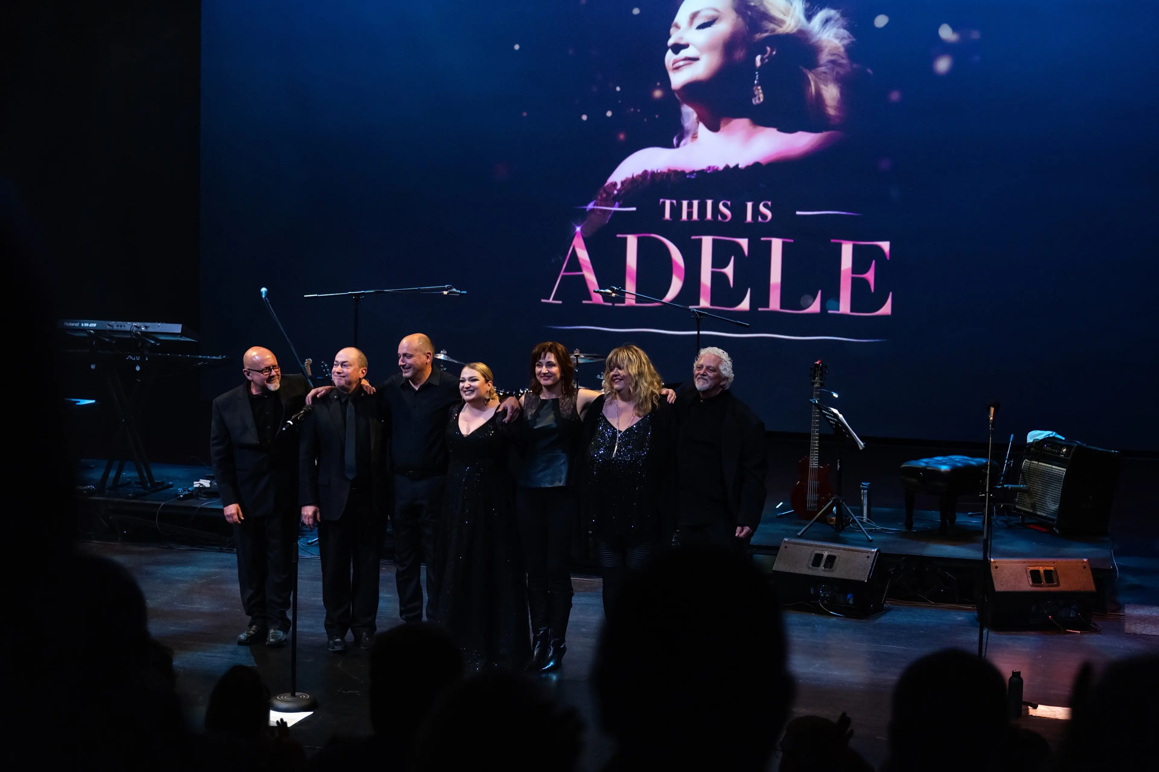 Six people standing arm in arm on stage with a large screen behind them displaying a woman's face and the text 'This is Adele'.