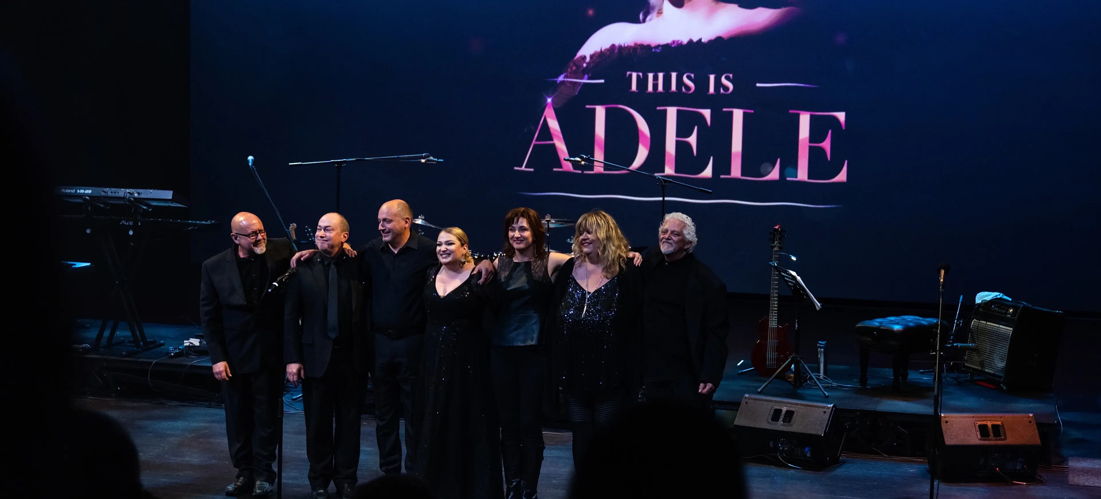 Six people standing arm in arm on stage with a large screen behind them displaying a woman's face and the text 'This is Adele'.