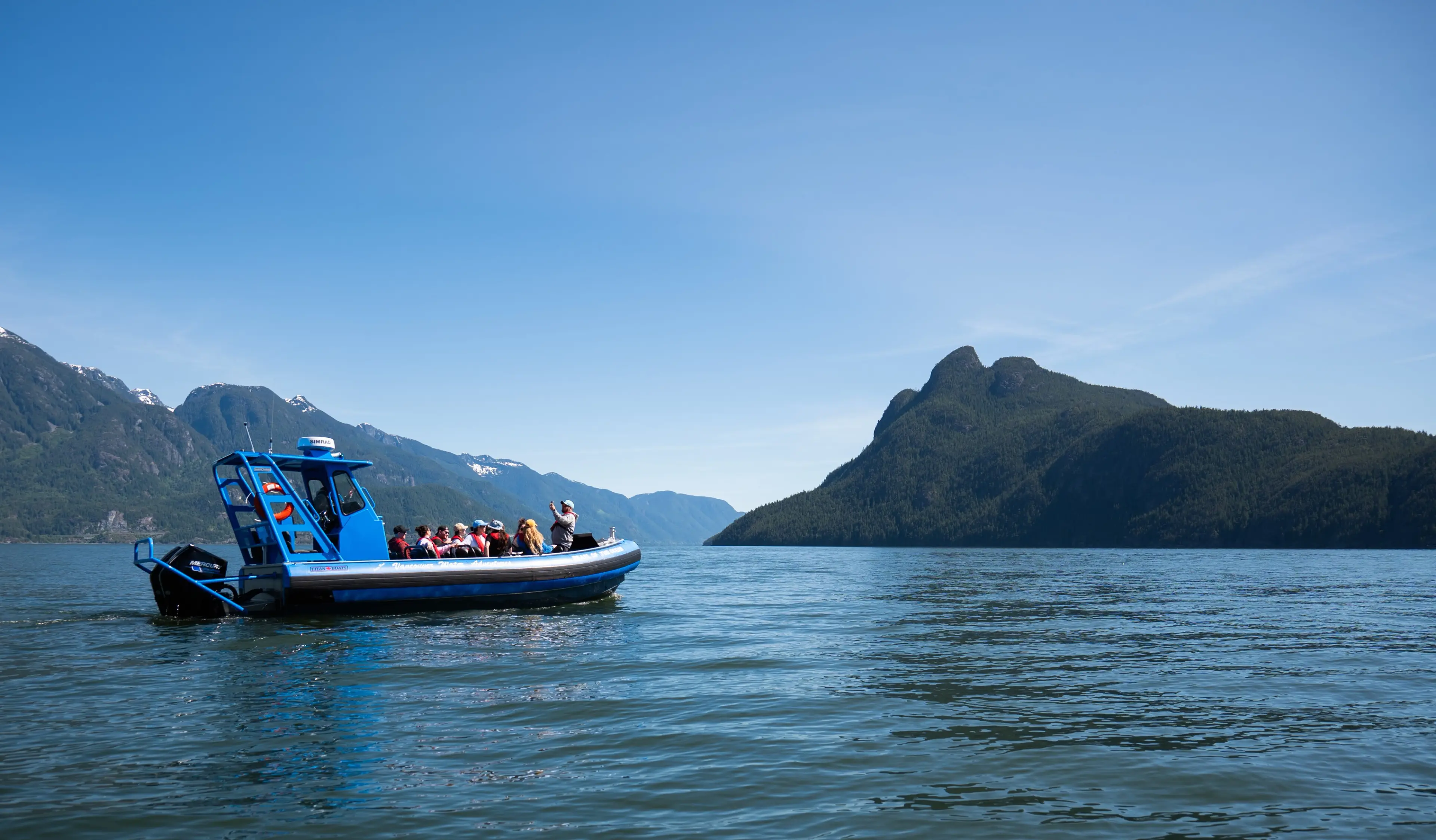 Zodiac boat with people wearing life jackets on calm water near forested mountains under clear sky.