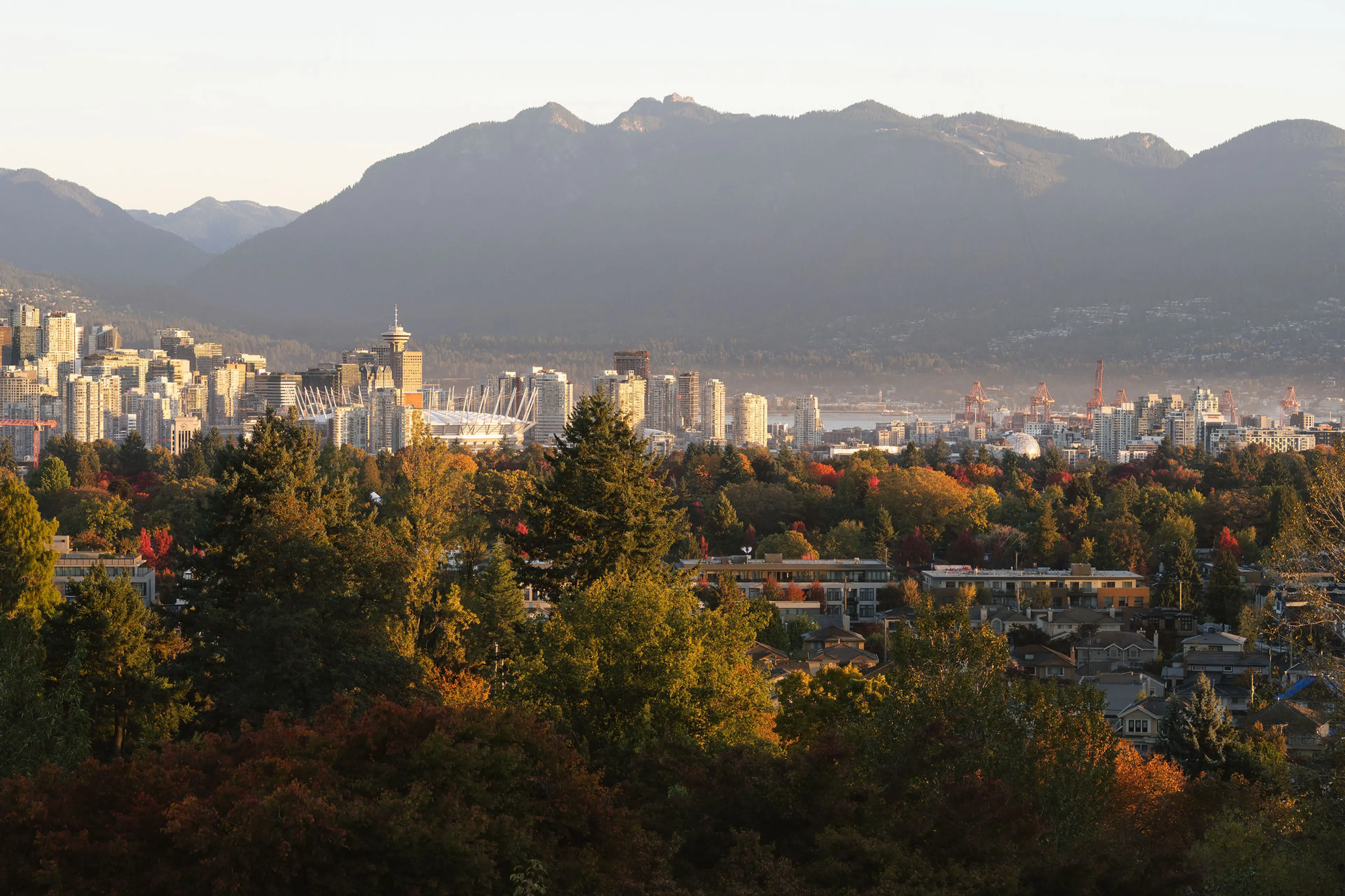 City skyline with tall buildings and stadium in front of forested area and mountains in the background during sunset.