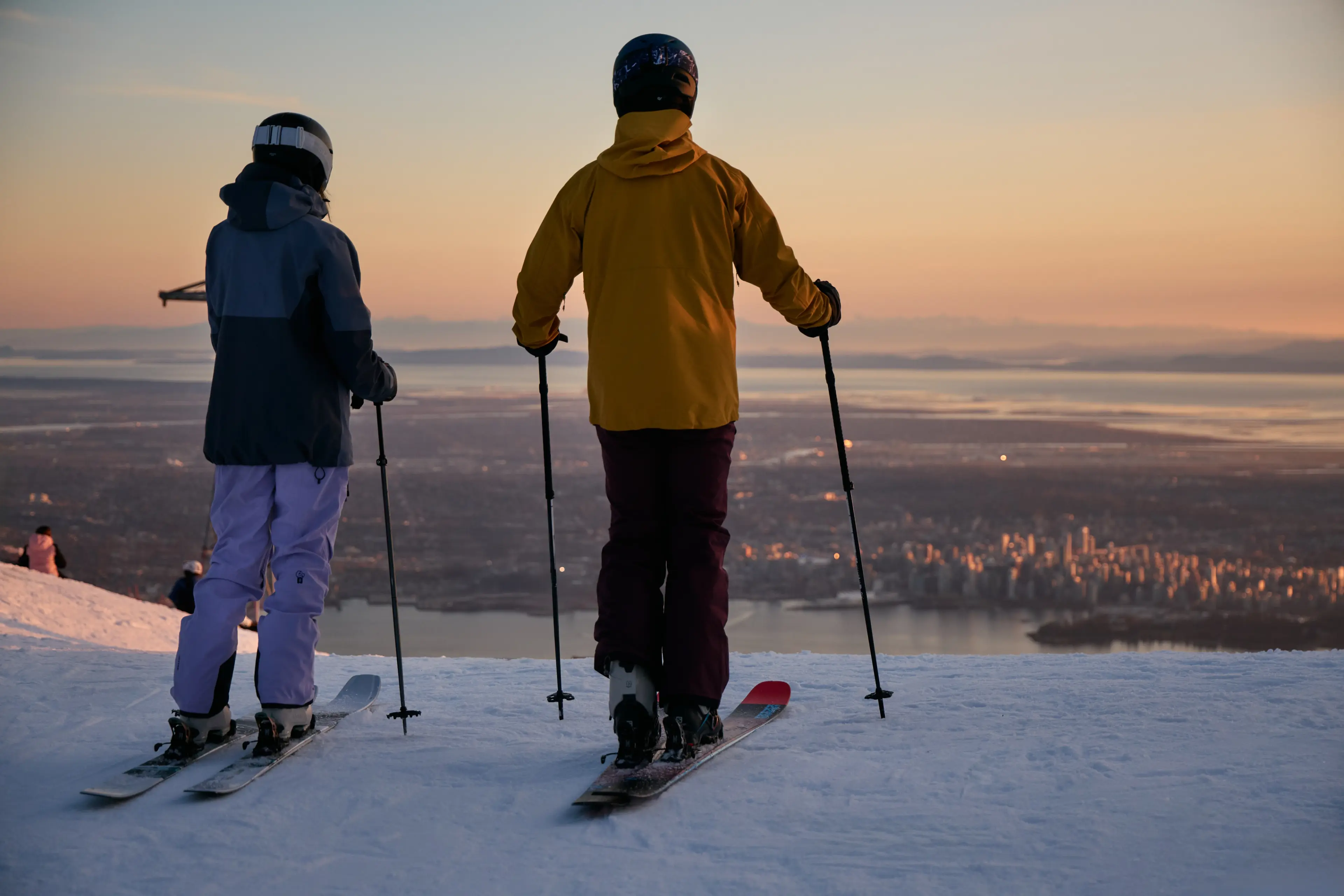 Two skiers in helmets and jackets standing on a snowy slope overlooking a city at sunset.
