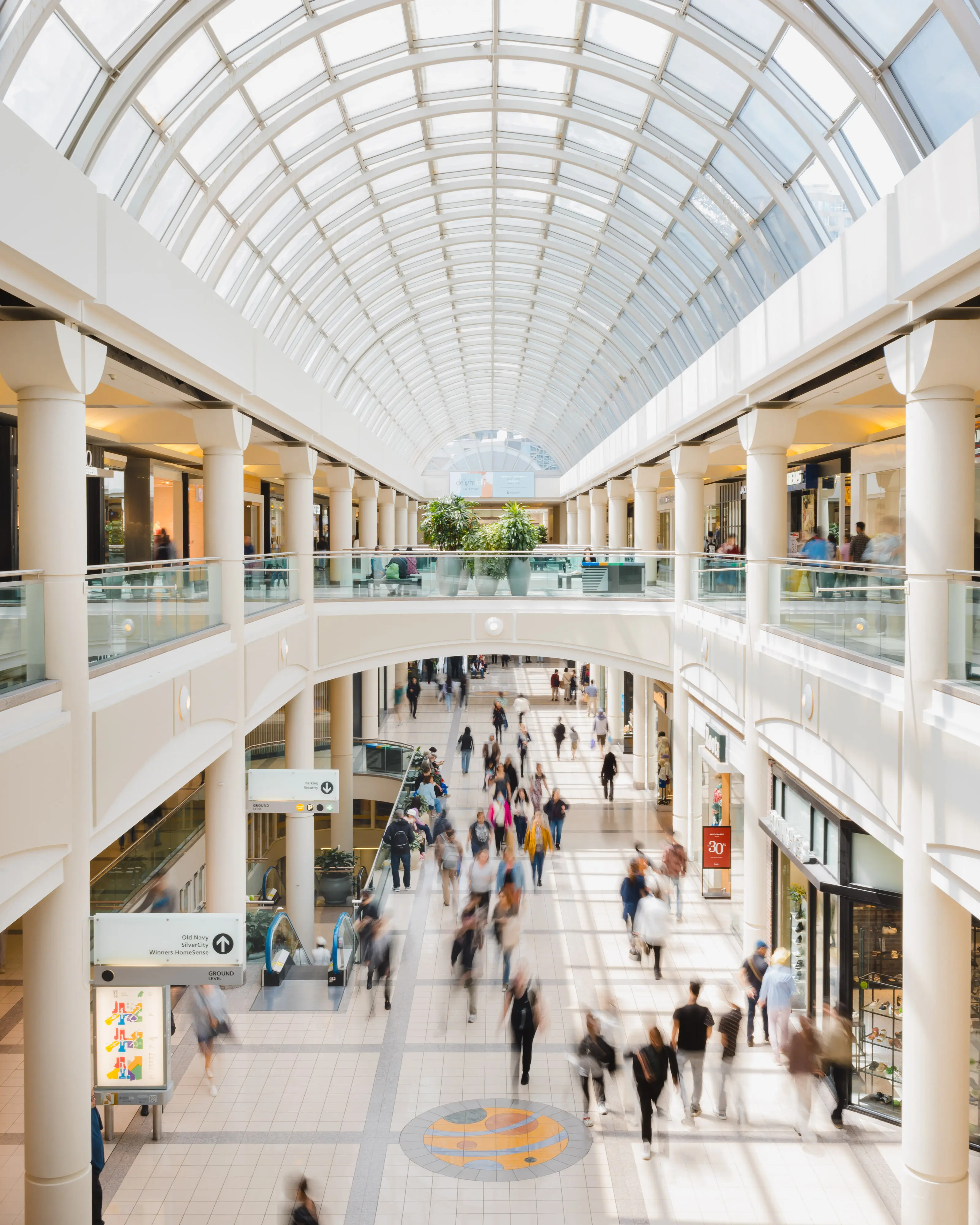 Busy shopping mall corridor with people walking under a large glass arched ceiling and white columns.