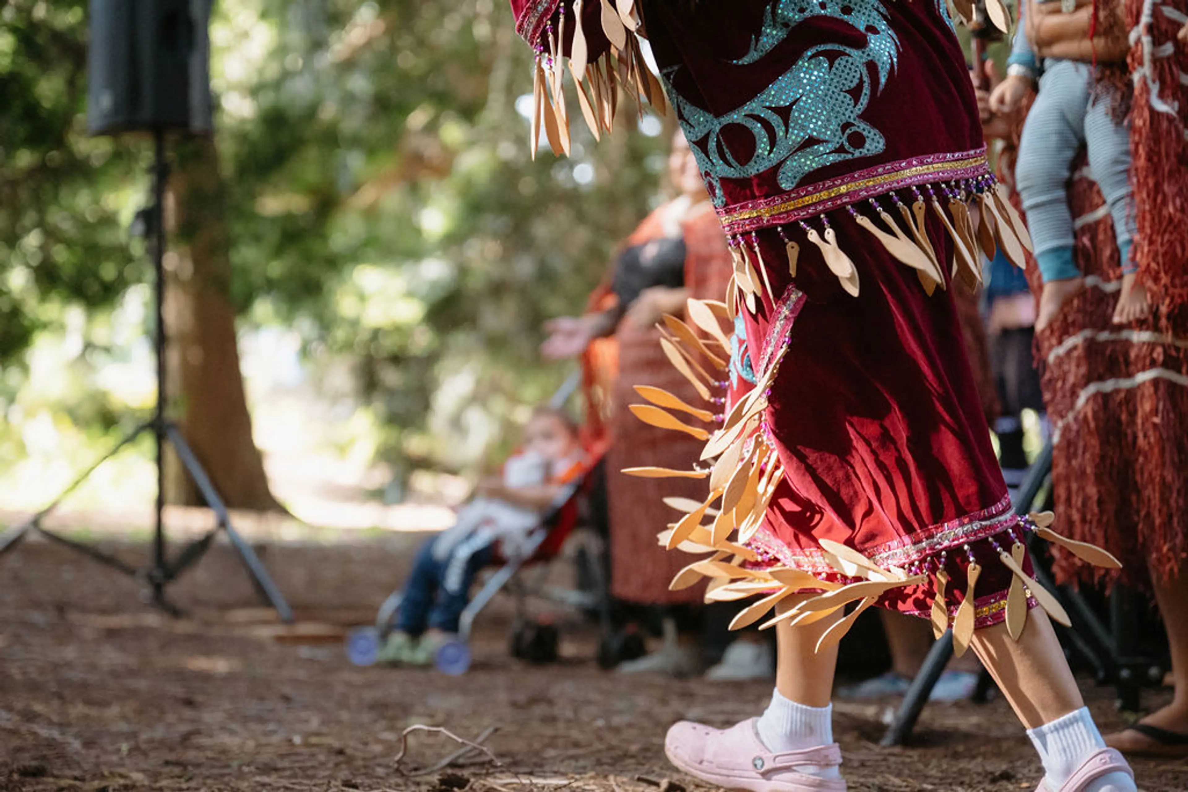 Person wearing a decorated maroon skirt with blue patterns and wooden ornaments, dancing outdoors on a dirt ground.