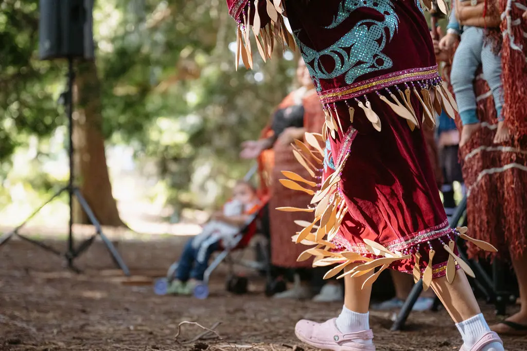 Person wearing a decorated maroon skirt with blue patterns and wooden ornaments, dancing outdoors on a dirt ground.