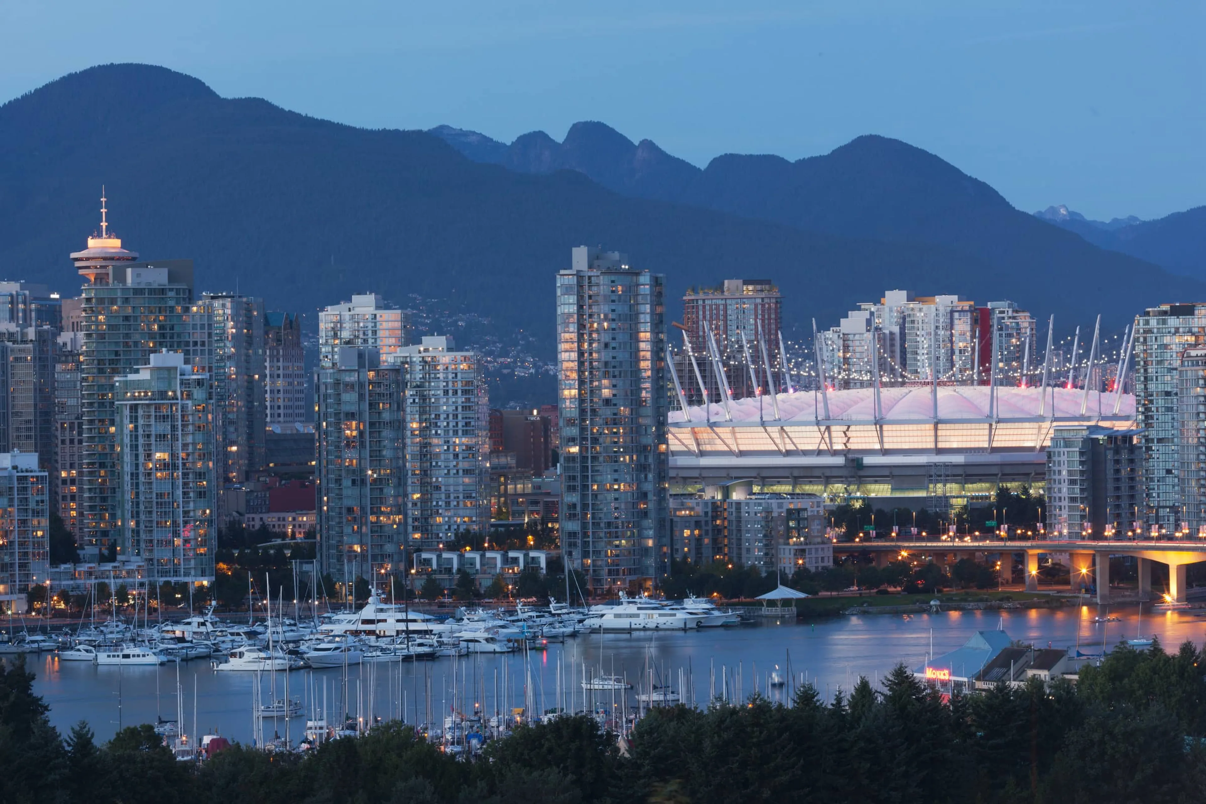 A breathtaking dusk view of the Vancouver skyline featuring the illuminated BC Place stadium and the Vancouver Lookout tower, with luxury yachts docked in False Creek against a backdrop of the North Shore Mountains.
