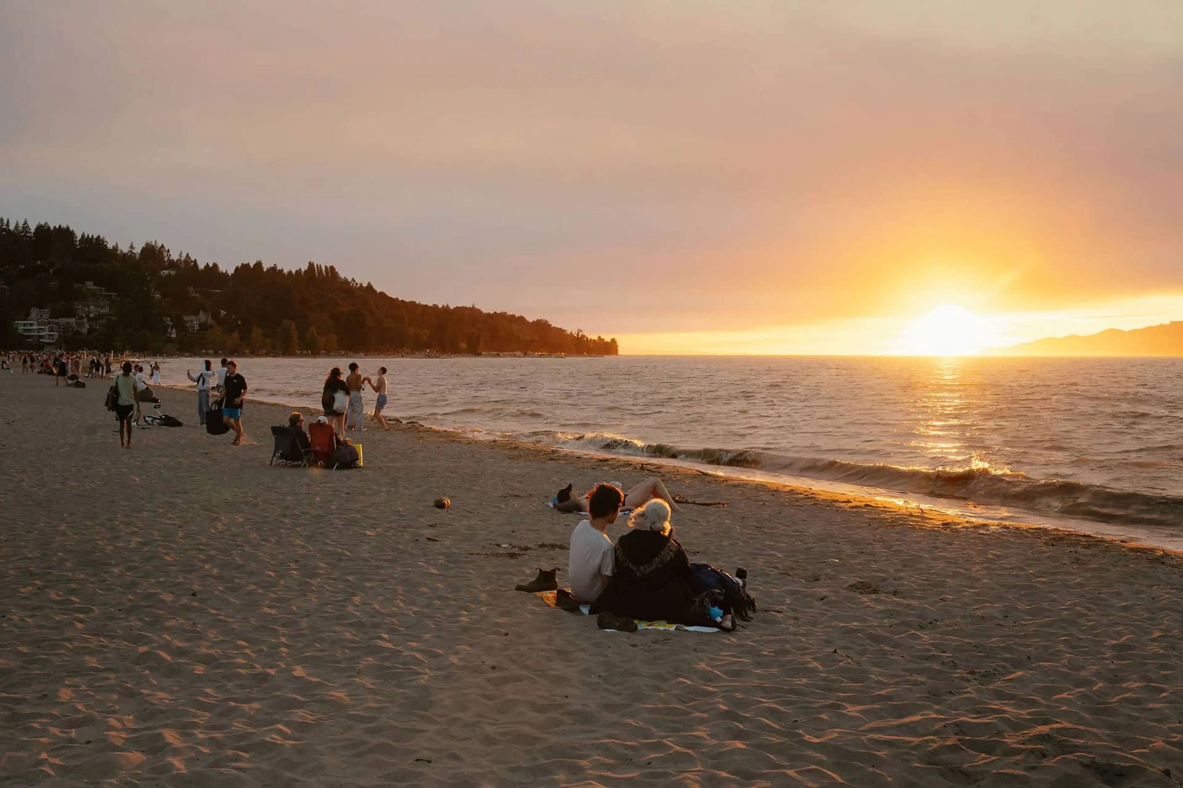 People sitting and walking on a sandy beach at sunset with the sun low over the ocean.