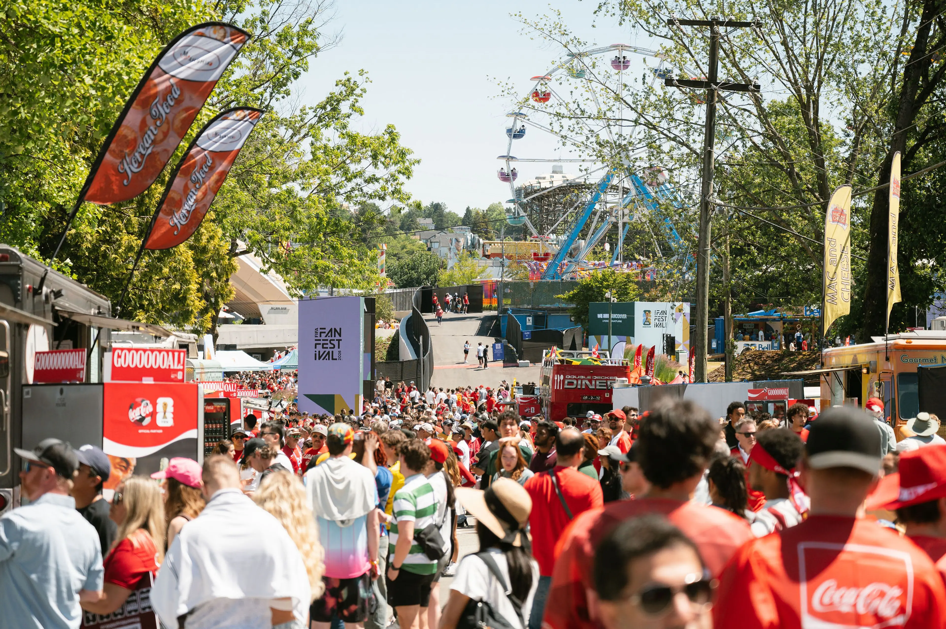 Crowd at an outdoor festival with food trucks, flags, and a Ferris wheel in the background on a sunny day.