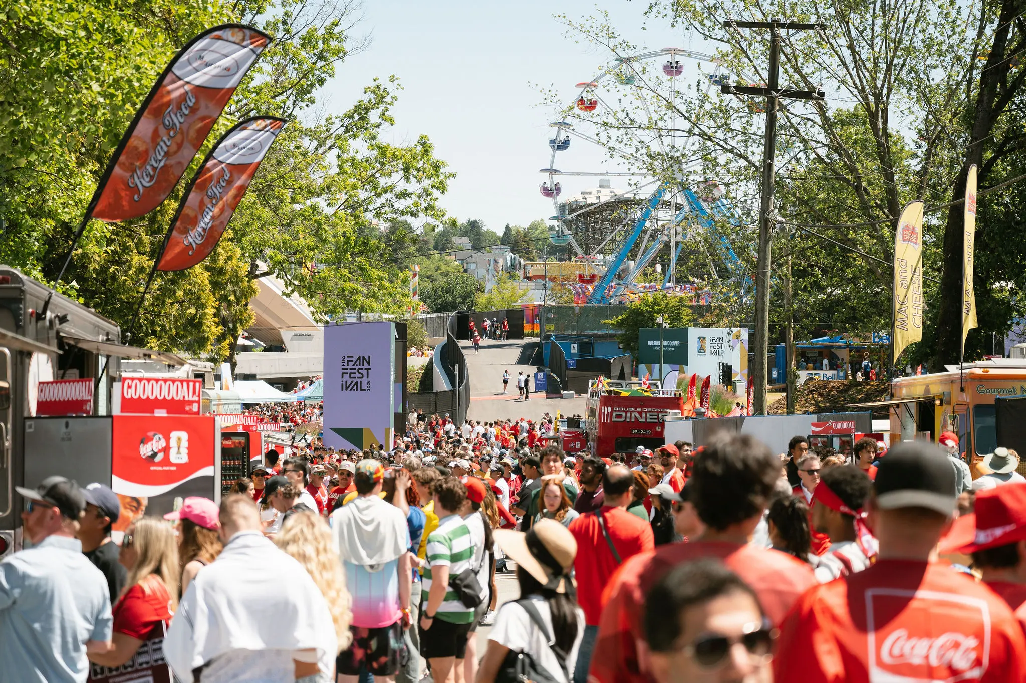 Crowd at an outdoor festival with food trucks, flags, and a Ferris wheel in the background on a sunny day.