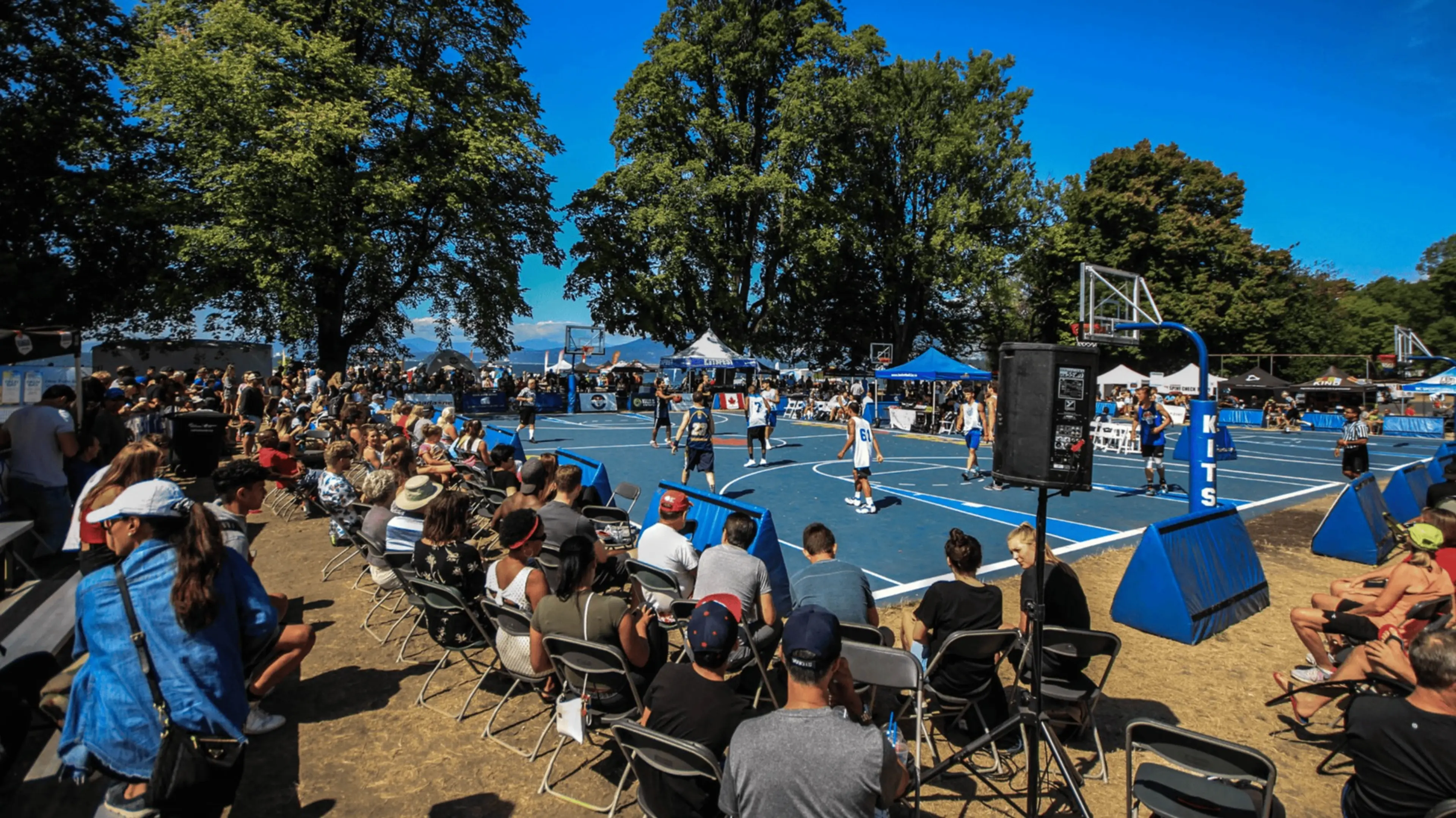 Outdoor basketball game on a blue court with players in white and dark jerseys and a crowd seated on folding chairs under trees.
