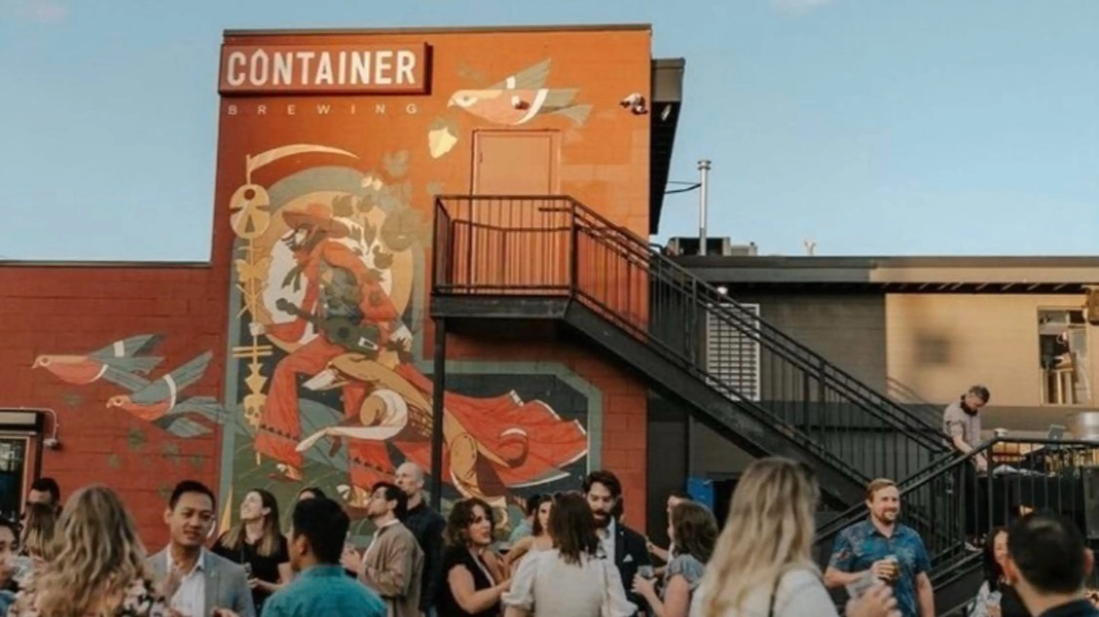 Crowd of people socializing outside a building with a mural and a sign reading Container Brewing.