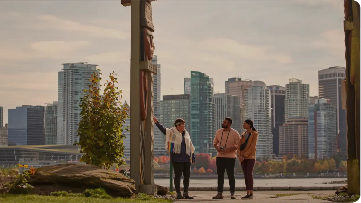 Three people stand by a tall totem pole in a park, with one pointing at it—an iconic stop on any Vancouver itinerary. Modern city skyscrapers and colorful autumn trees brighten the background under a partly cloudy sky.