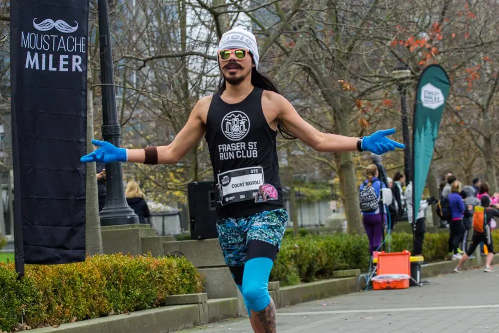 Runner wearing a white beanie, sunglasses, blue gloves, and colorful shorts with a black tank top that says Fraser St Run Club, running with arms outstretched near a Moustache Miler banner.