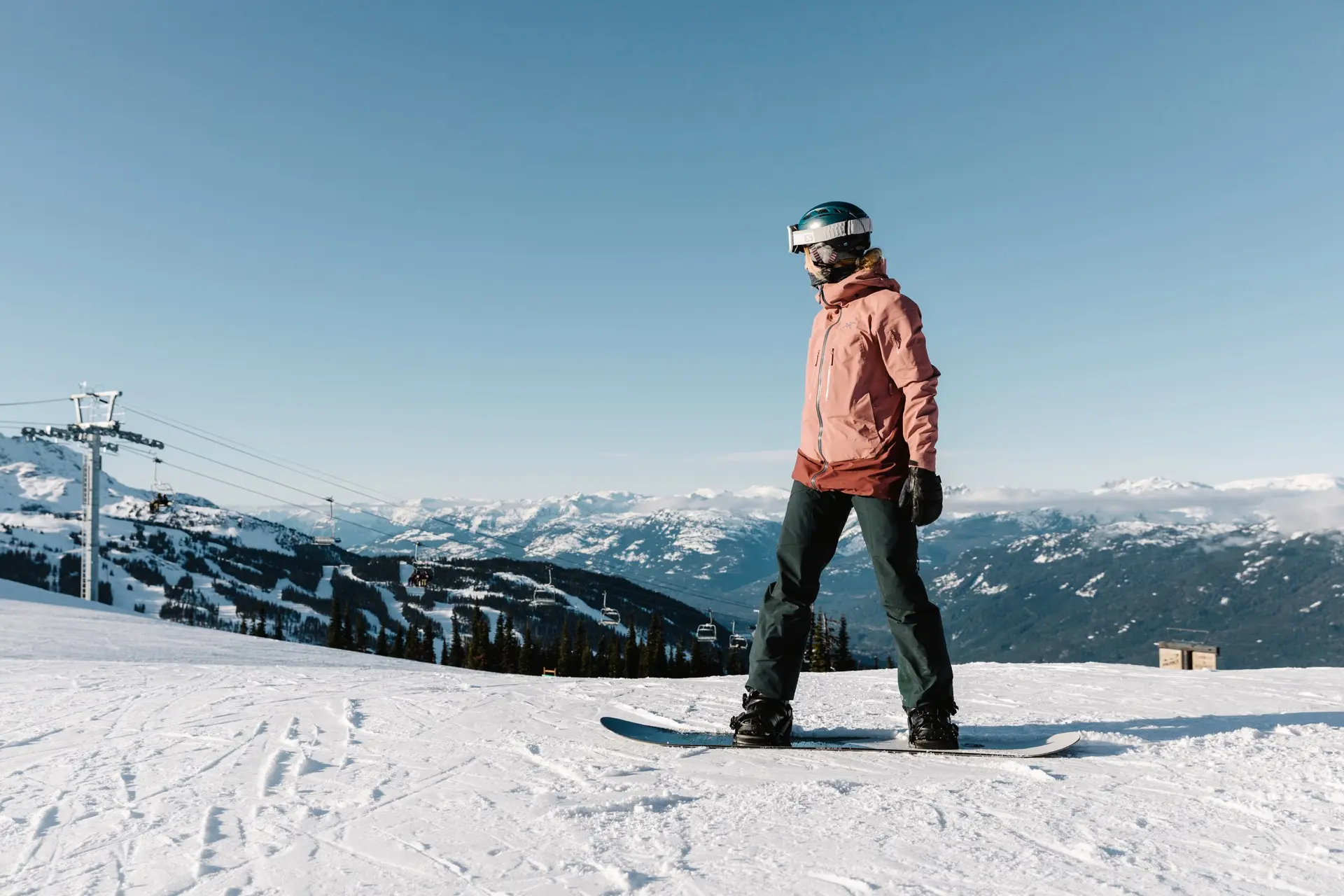 A person wearing a helmet and goggles stands on a snowboard atop a snowy mountain, with ski lifts and distant mountains visible under a clear blue sky—perfect for adventurous day trips from Vancouver.