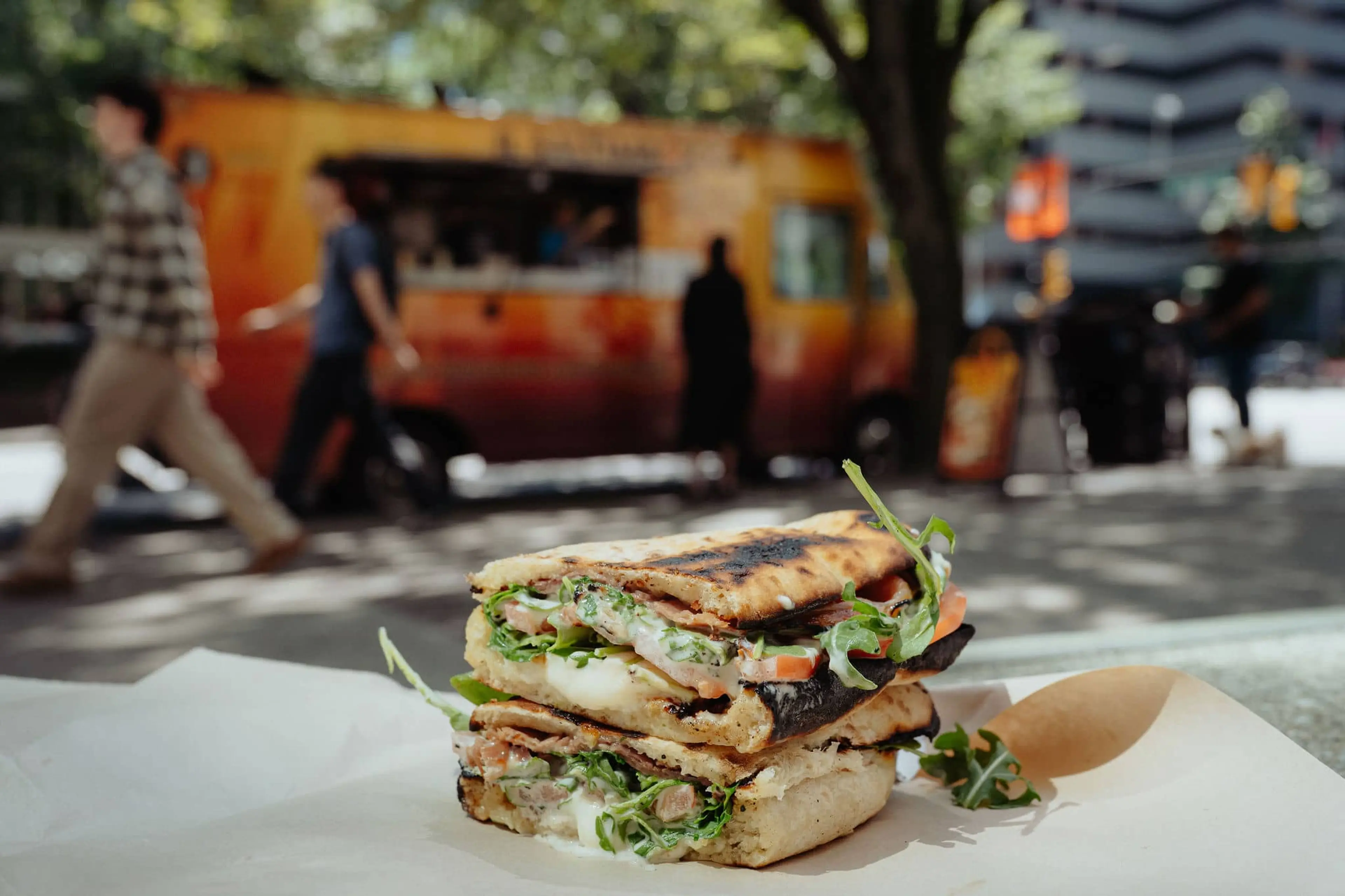 Grilled sandwich with greens, tomatoes, and cheese on white paper with blurred food truck and people in background
