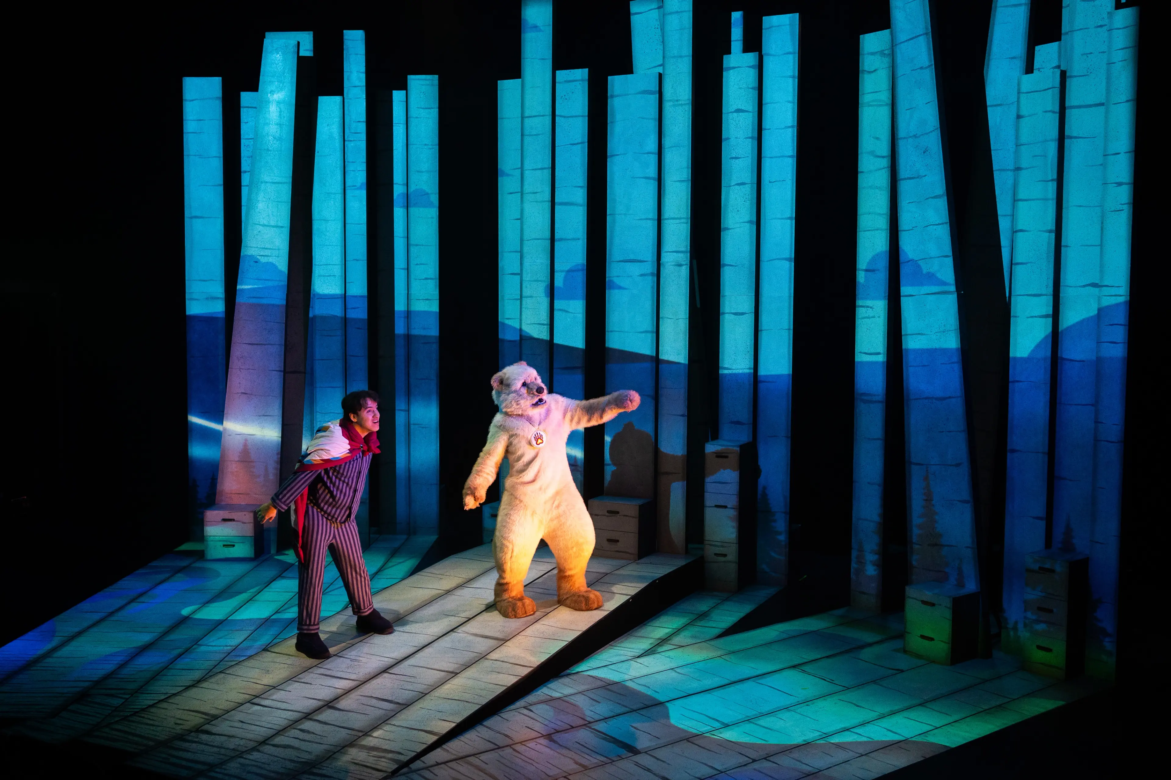 A person in striped pajamas and a red scarf stands next to a person in a bear costume on a stage with tall blue pillars and forest projections.