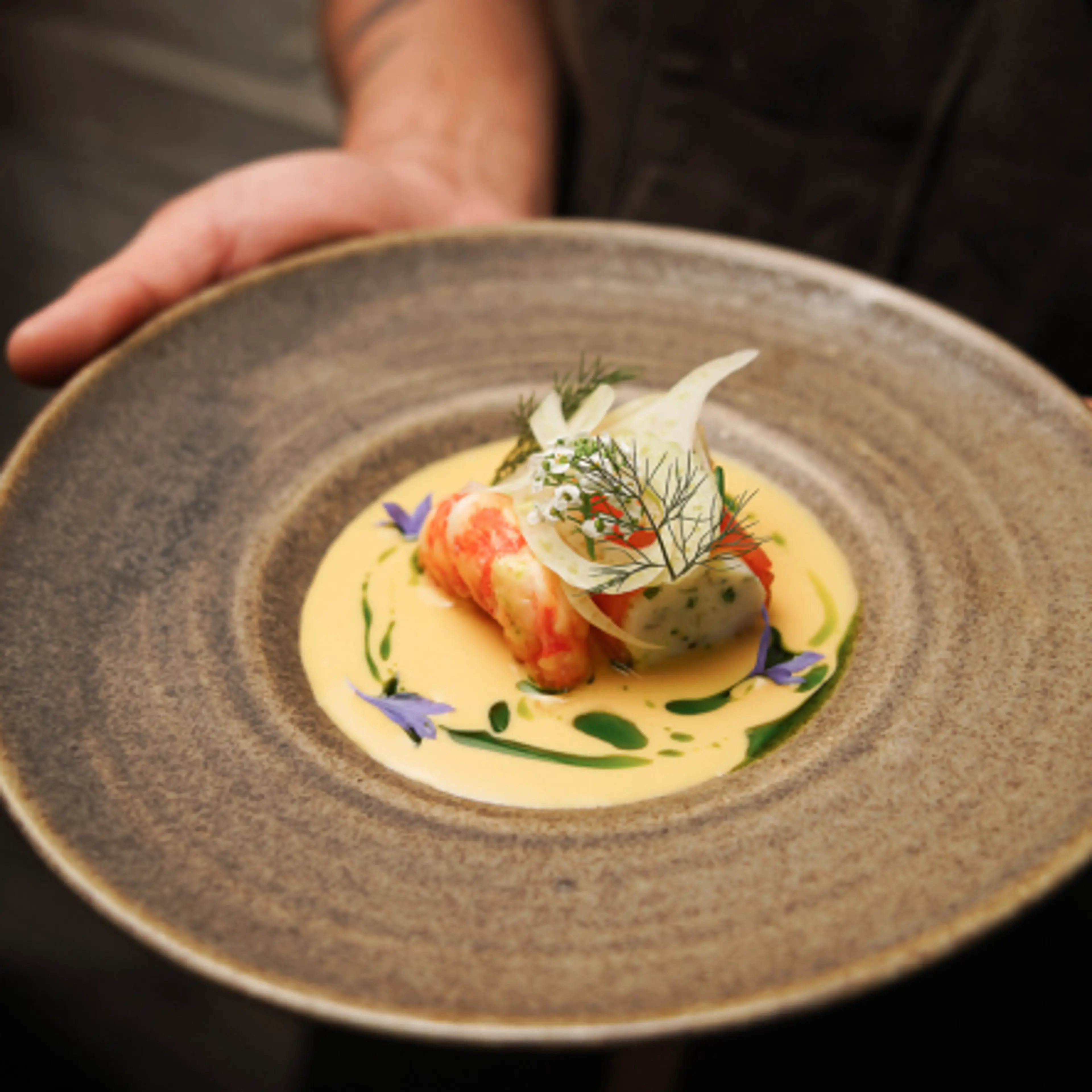 Hand holding a rustic plate with a gourmet dish featuring seafood, creamy sauce, herbs, and edible flowers.