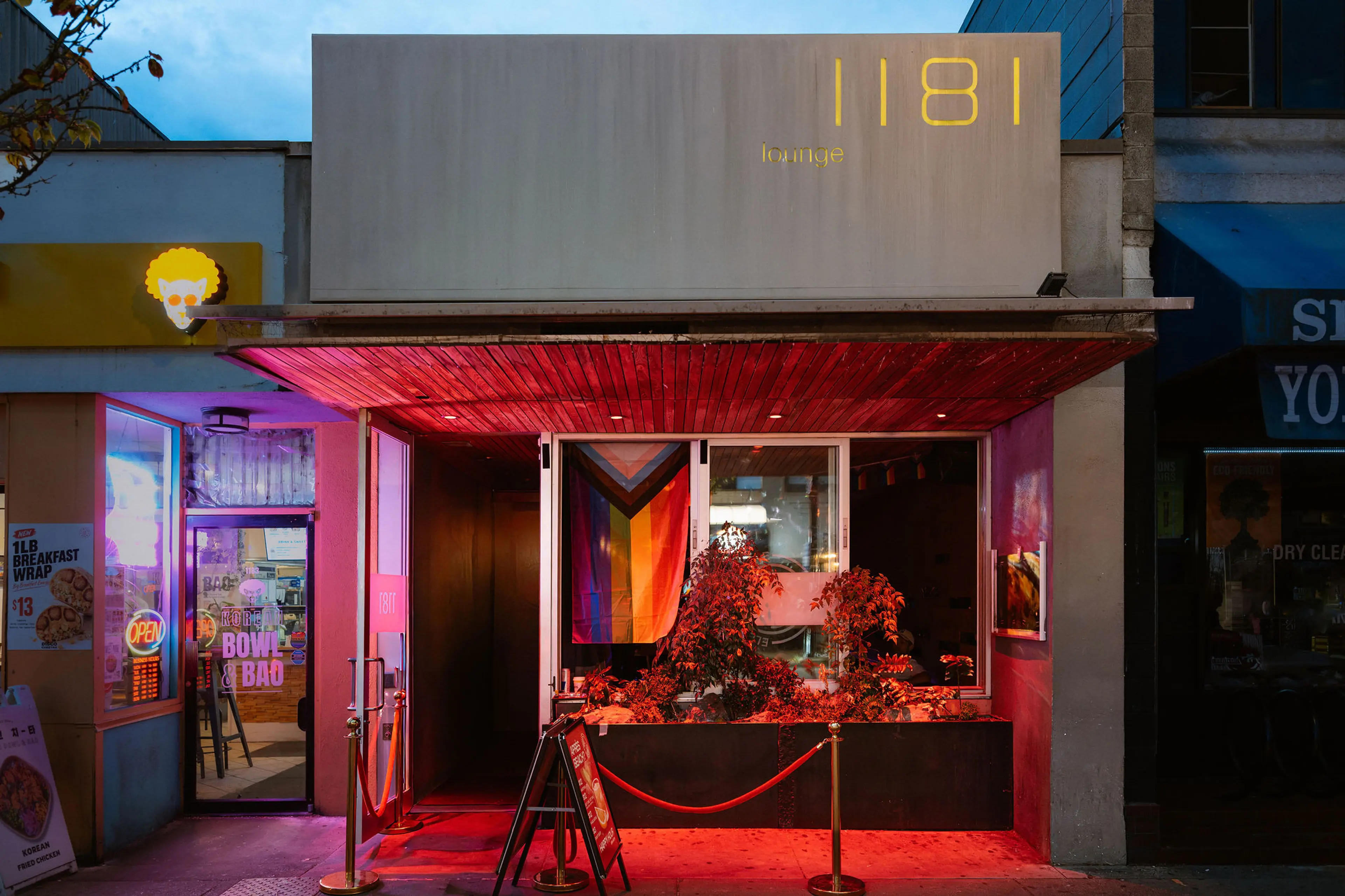 Entrance of 1181 lounge with red lighting, plants in front, and a colorful flag inside the window.