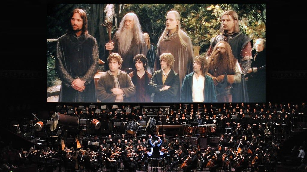 Orchestra performing live in front of a large screen showing a scene from The Lord of the Rings with characters standing in a forest.
