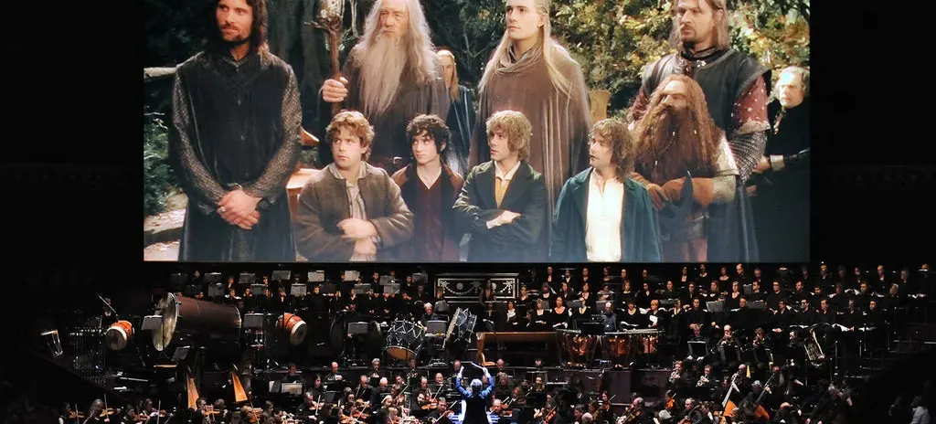 Orchestra performing live in front of a large screen showing a scene from The Lord of the Rings with characters standing in a forest.