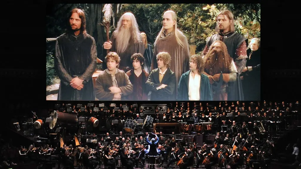 Orchestra performing live in front of a large screen showing a scene from The Lord of the Rings with characters standing in a forest.