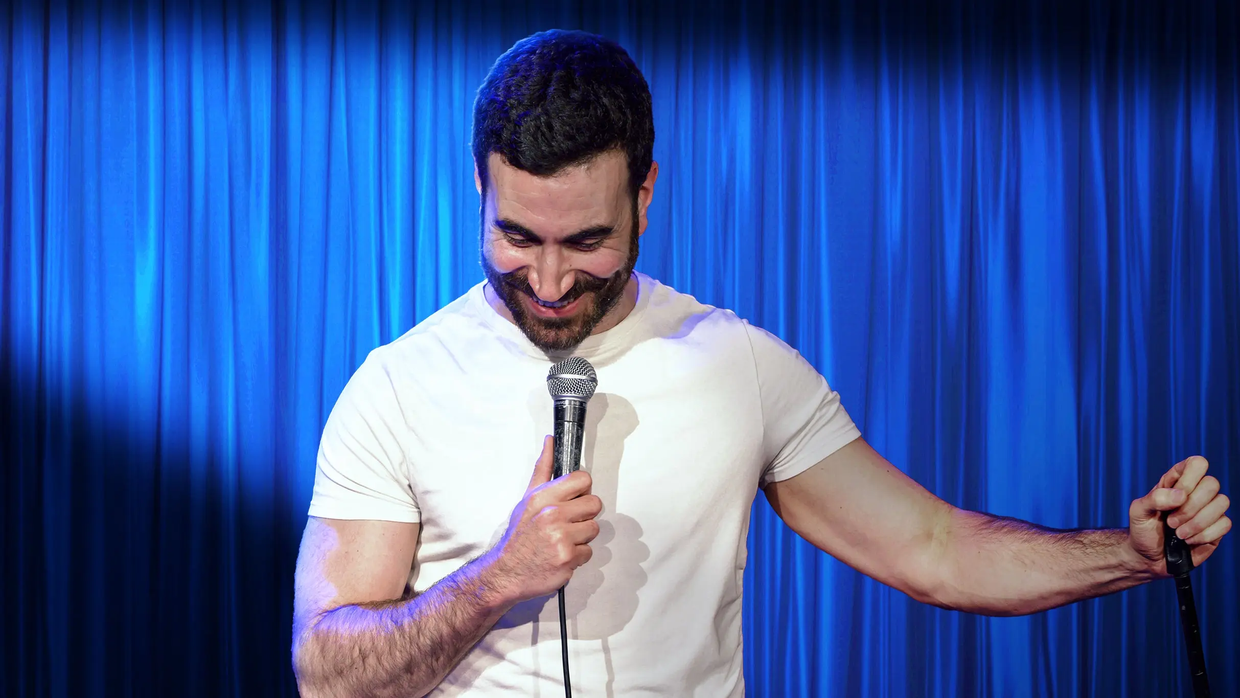 A person in a white t-shirt holding a microphone and smiling on a stage with blue curtains.