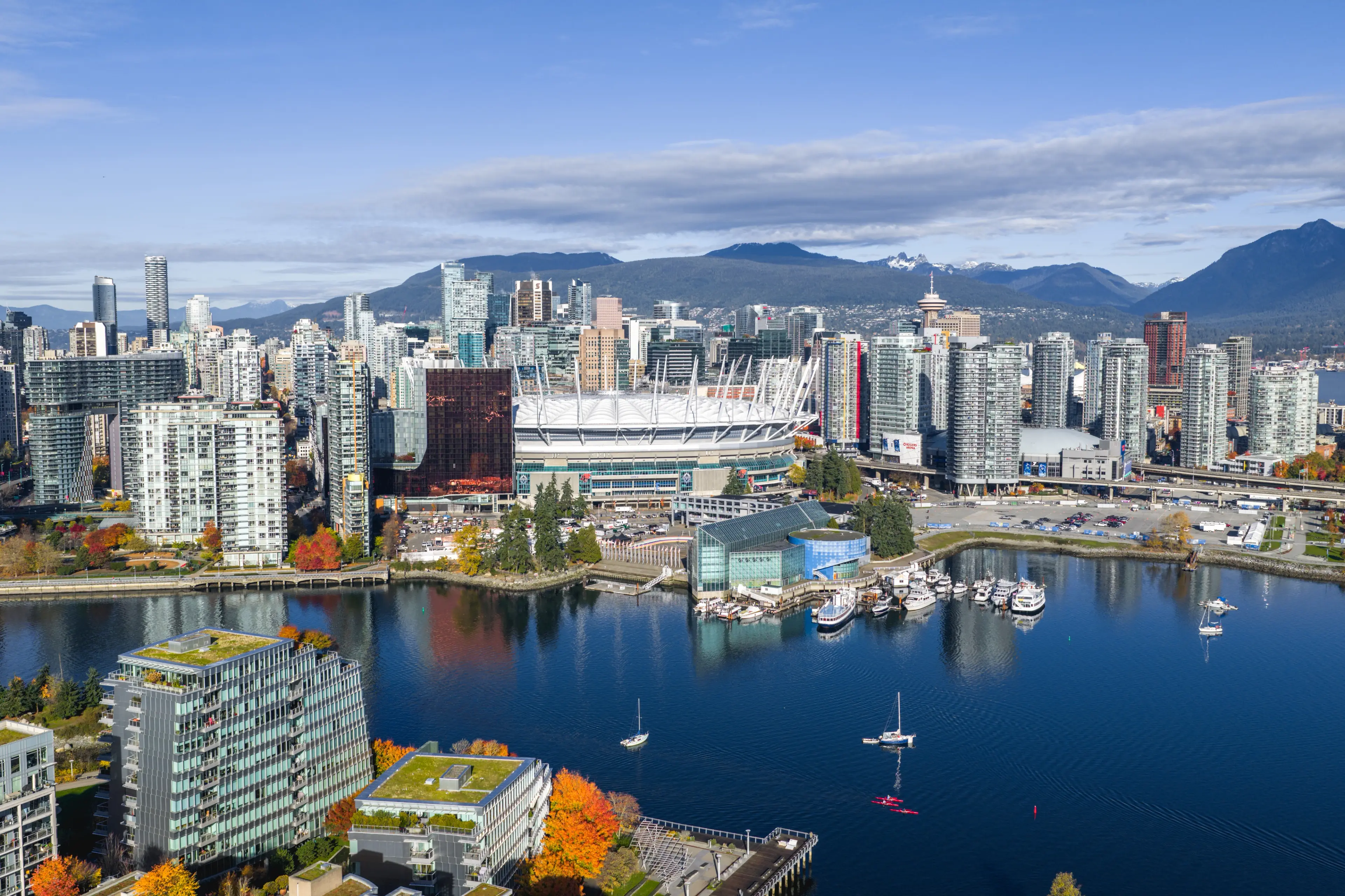 City skyline with modern high-rise buildings, BC Place stadium, boats on water, and mountains in the background under a partly cloudy sky.