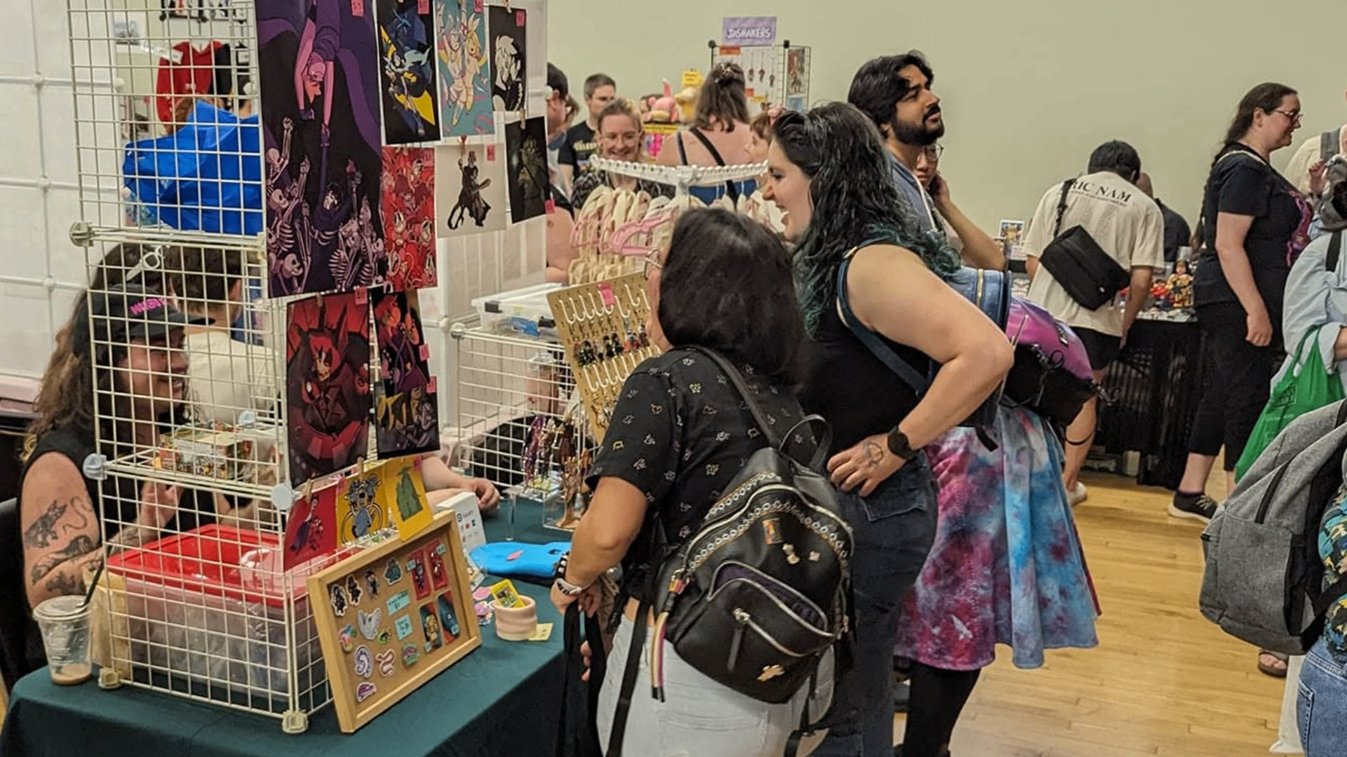 People browsing and shopping at a convention booth displaying artwork, pins, and jewelry on a table and grid panels.