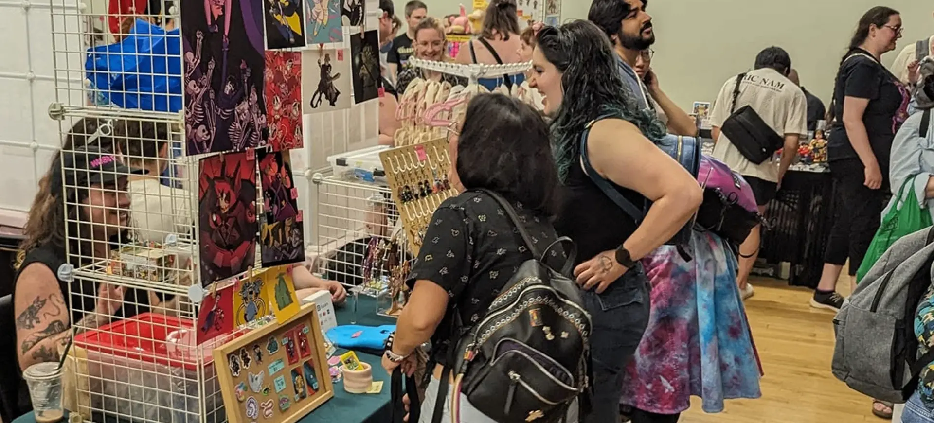 People browsing and shopping at a convention booth displaying artwork, pins, and jewelry on a table and grid panels.