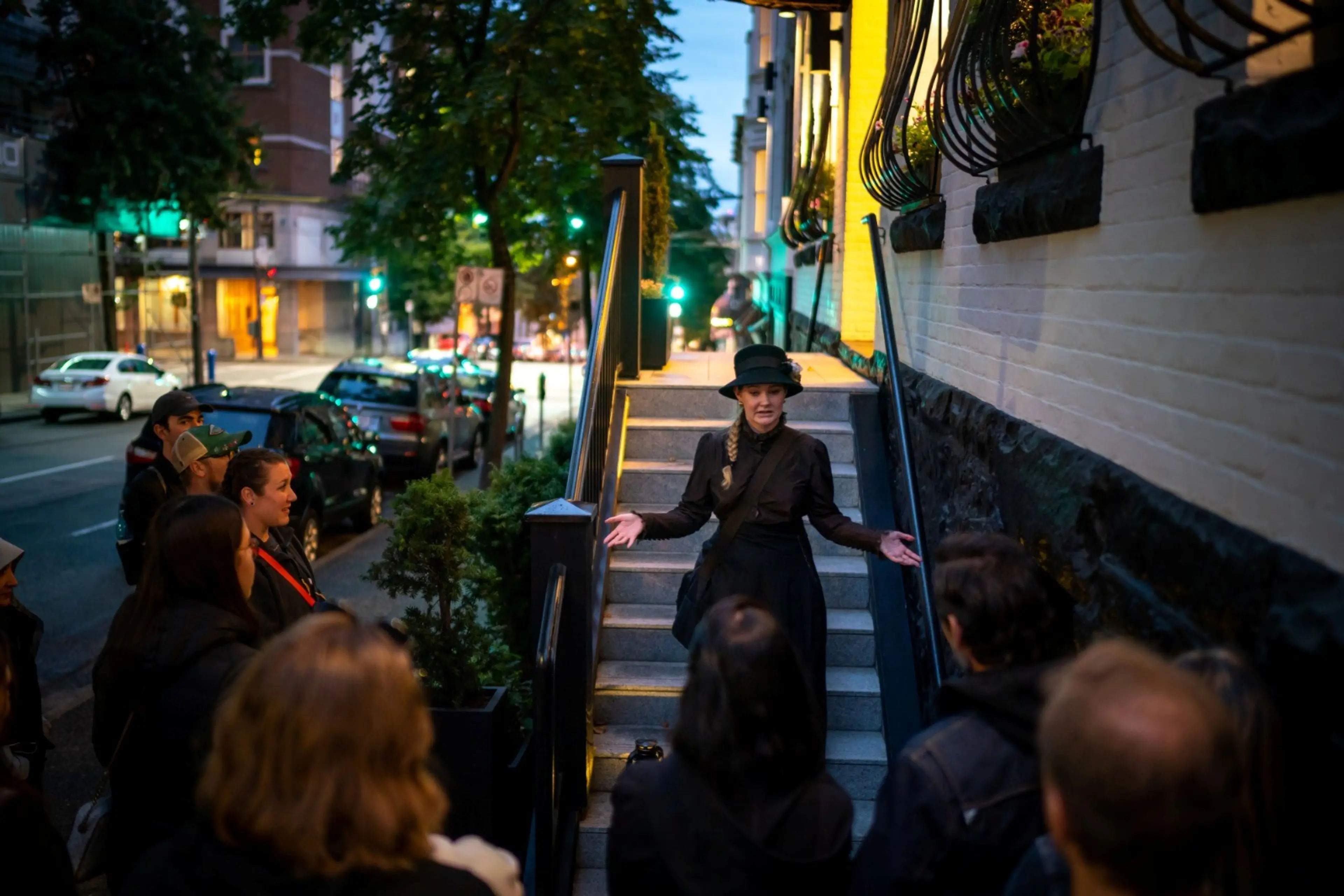 Woman with a hat and dress speaking to a group on a city sidewalk near stairs at dusk.