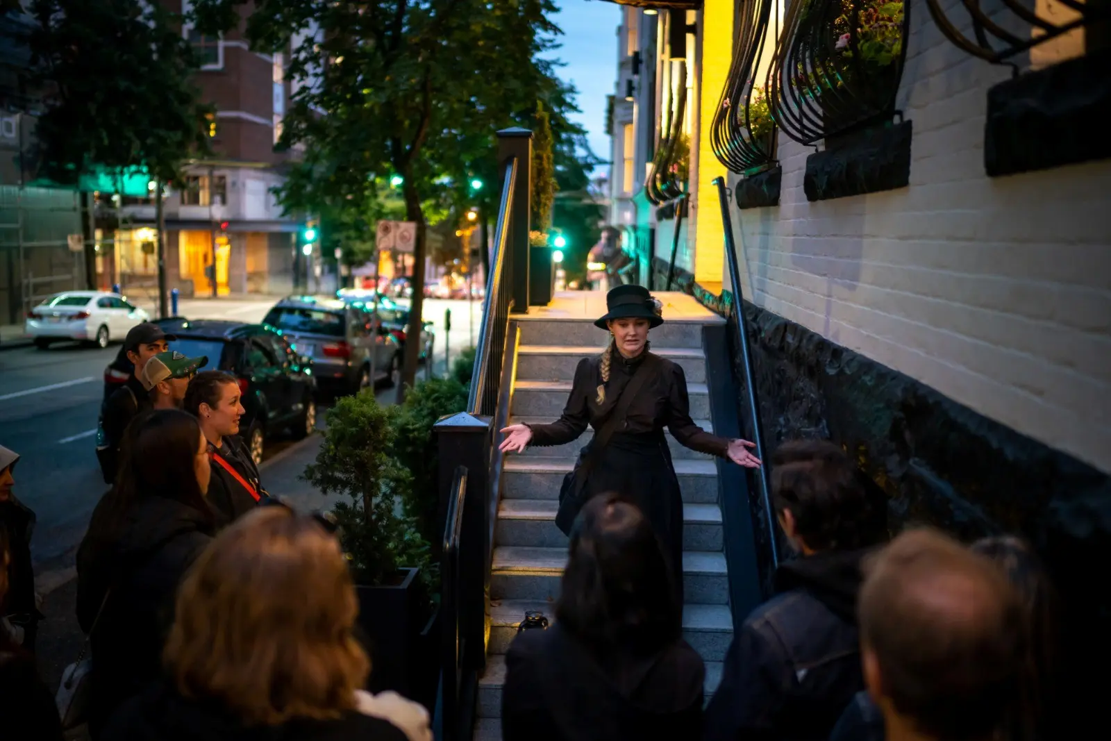 Woman with a hat and dress speaking to a group on a city sidewalk near stairs at dusk.