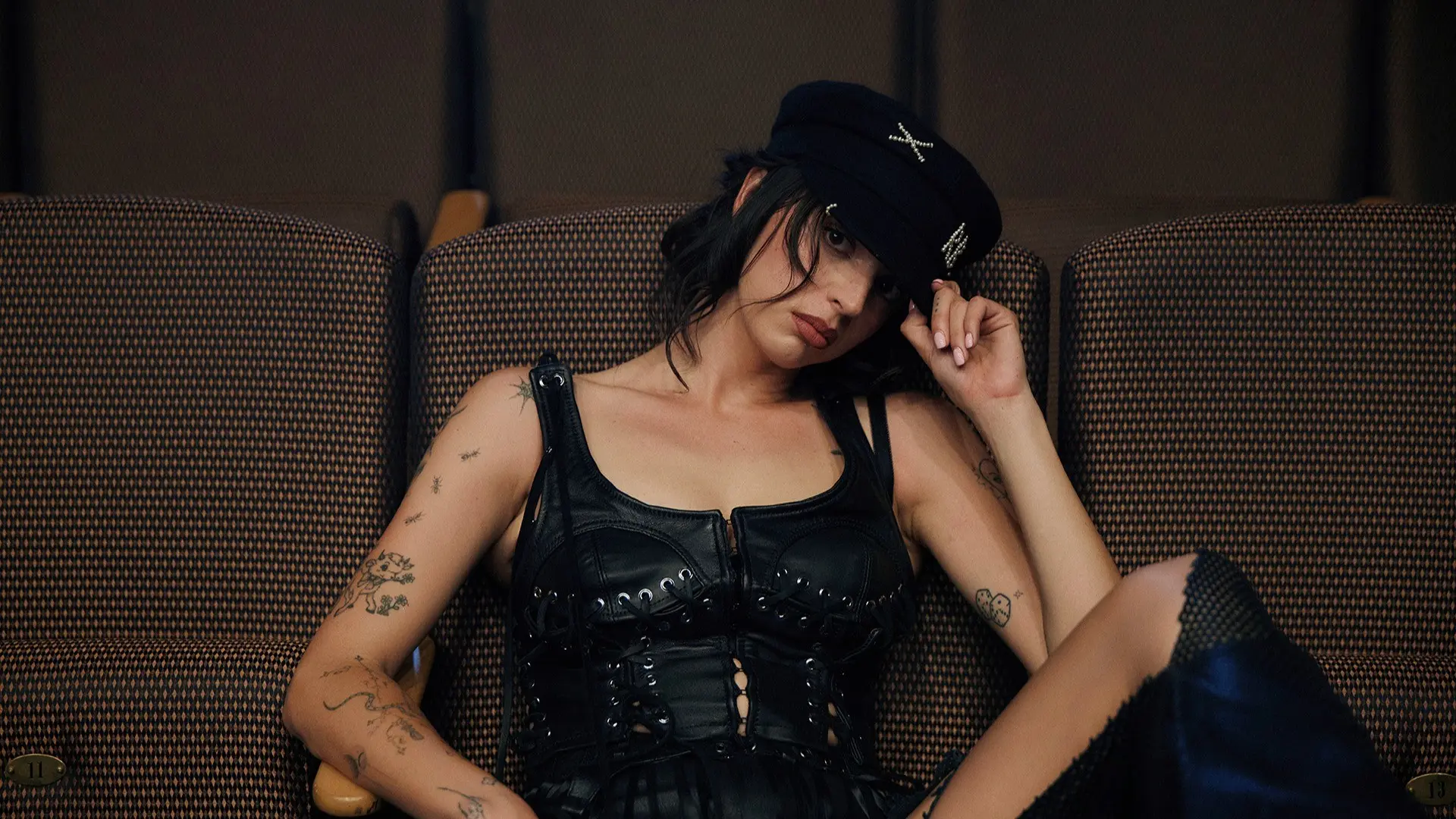 Person with tattoos wearing a black leather corset and black hat sitting on a patterned couch, tilting head and touching hat.