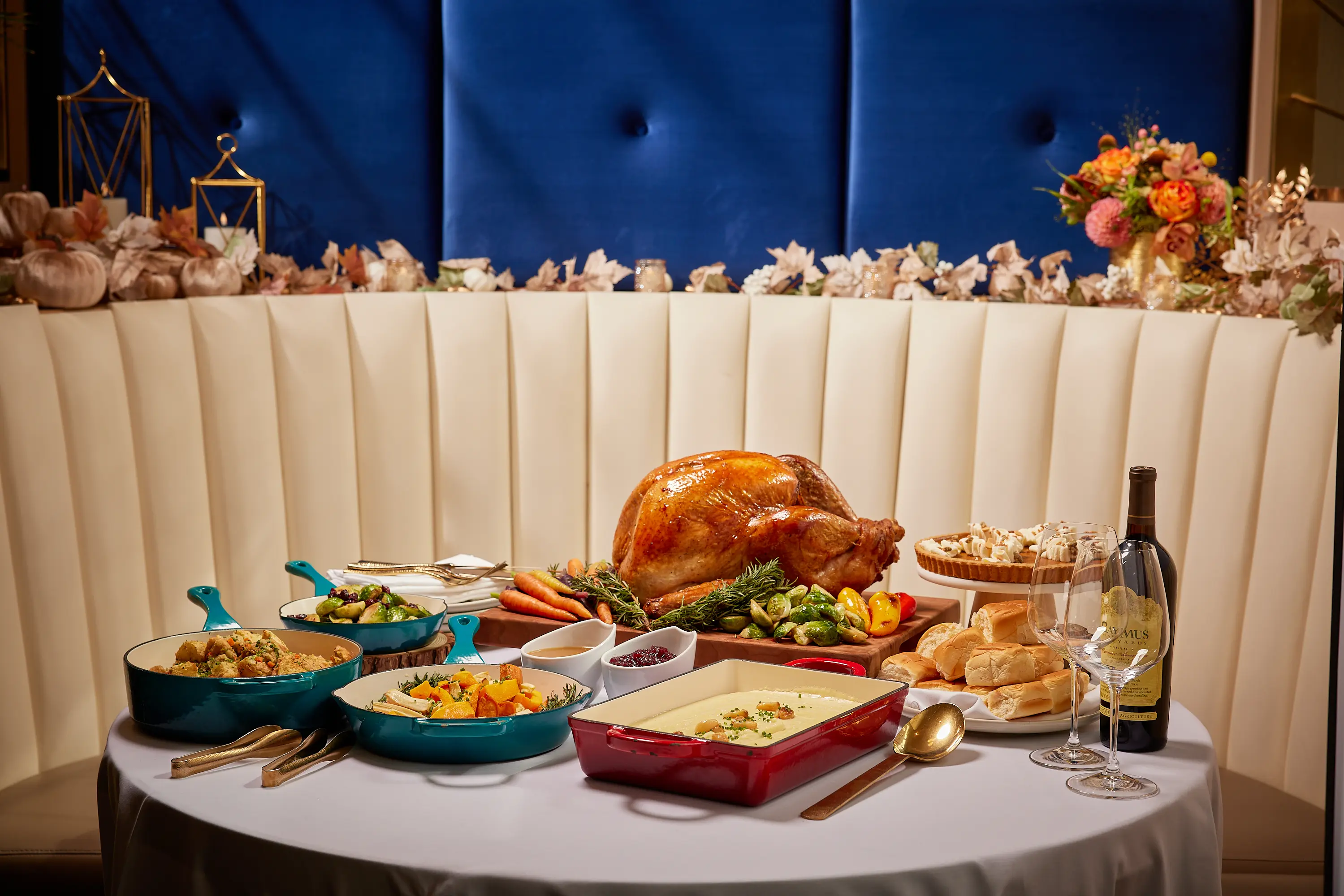Table with roasted turkey, vegetables, mashed potatoes, bread rolls, wine bottle, wine glasses, and pumpkin pie with whipped cream.
