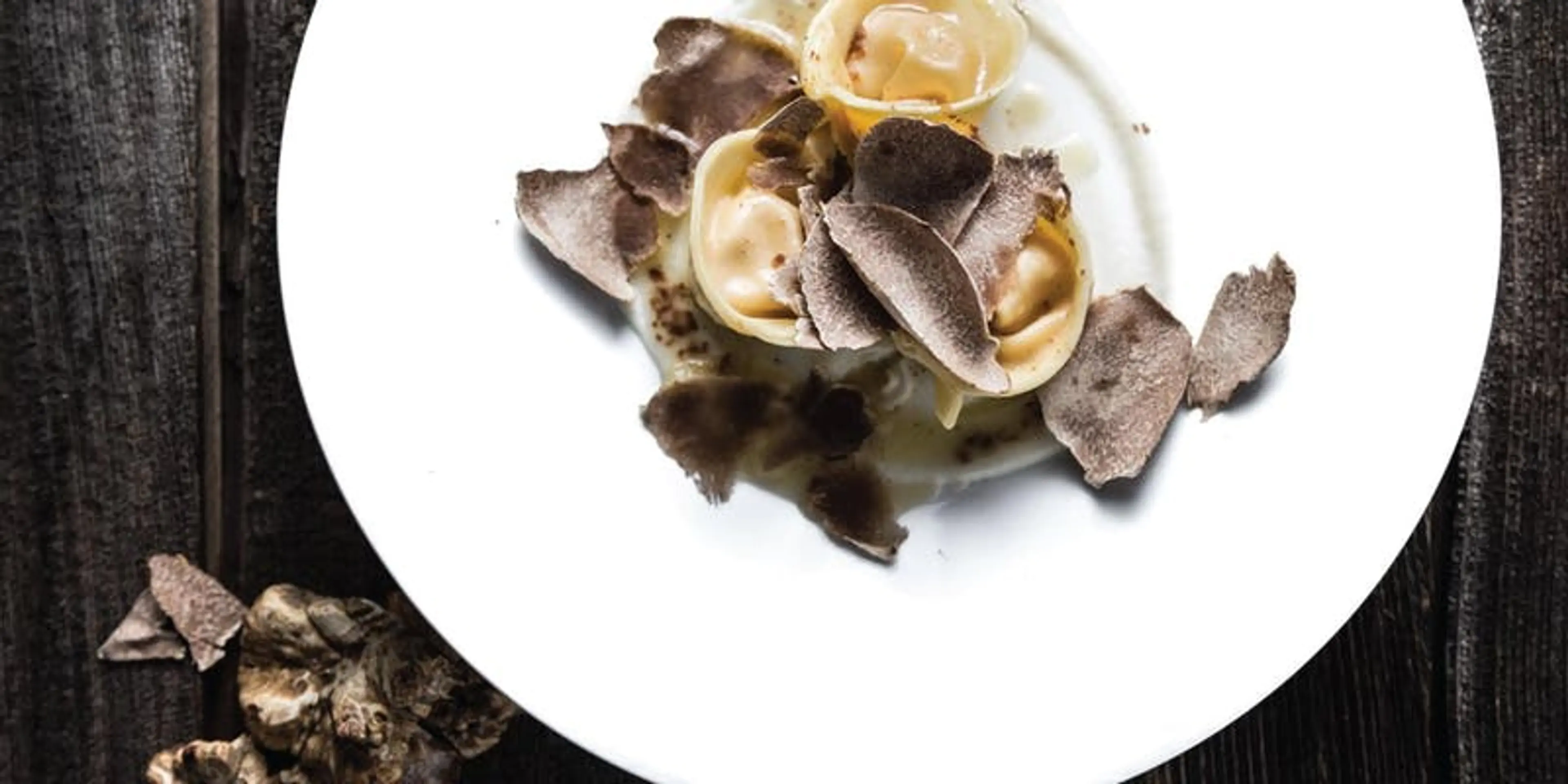 Three pieces of pasta with creamy sauce topped with shaved black truffles on a white plate over a dark wooden surface.