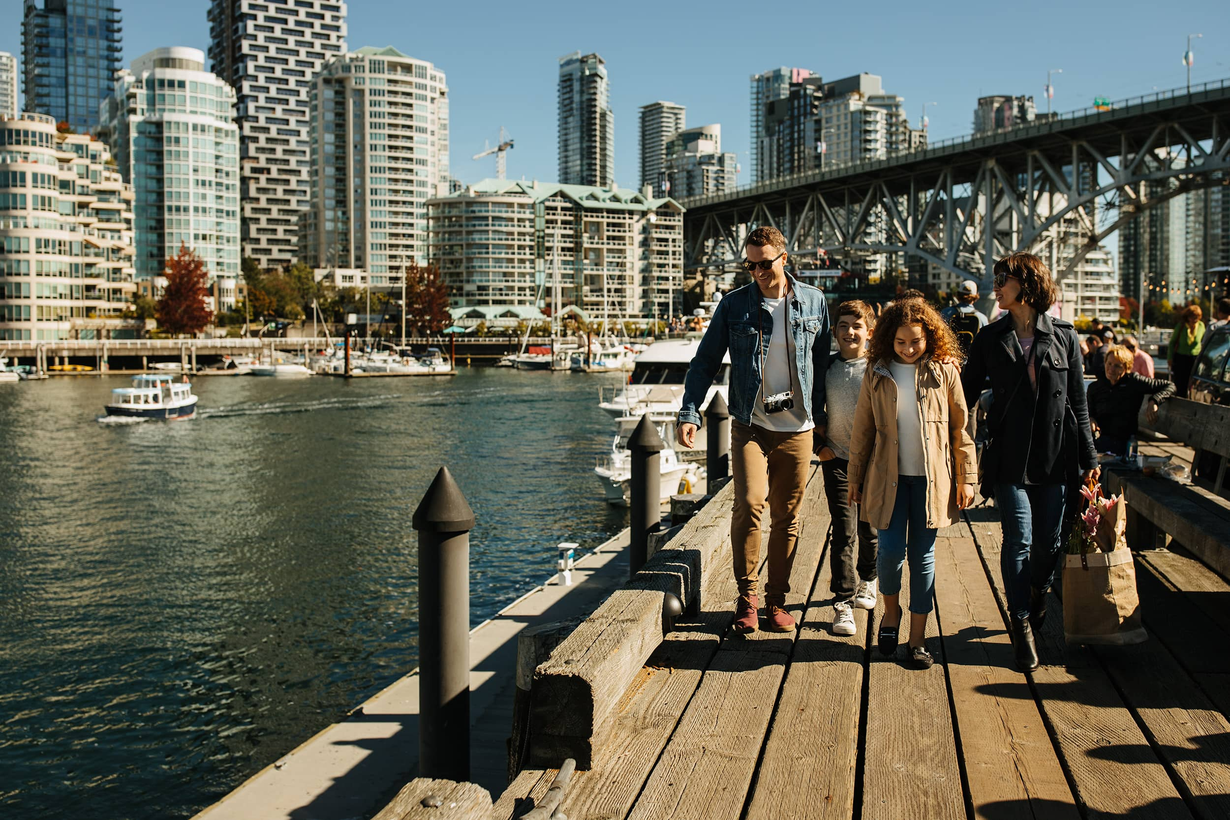 Family of four walking on a wooden pier by the water with city buildings and a bridge in the background.