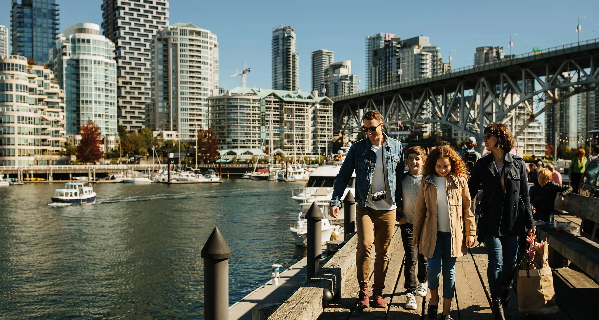 Family of four walking on a wooden pier by the water with city buildings and a bridge in the background.