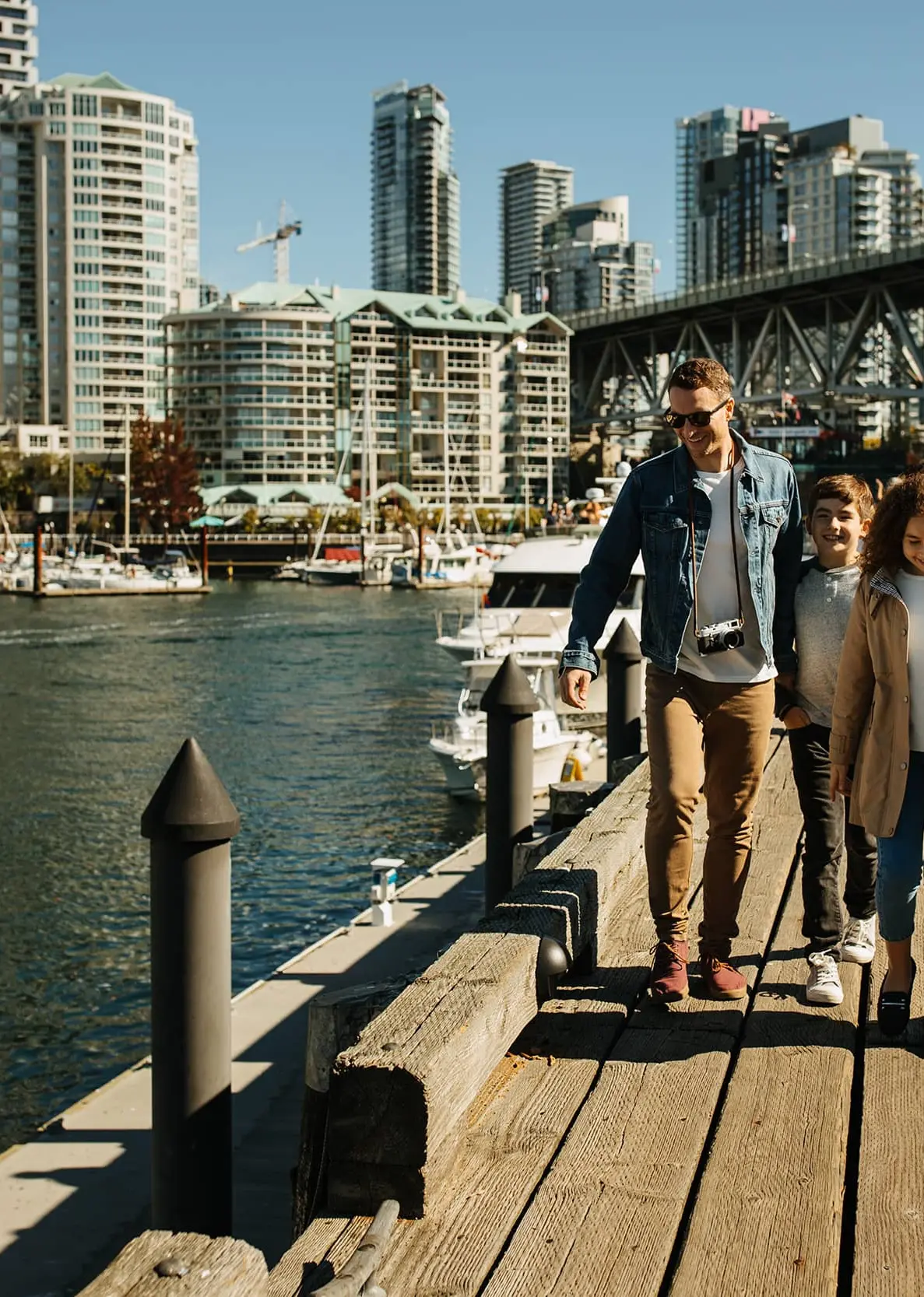 Family of four walking on a wooden pier by the water with city buildings and a bridge in the background.