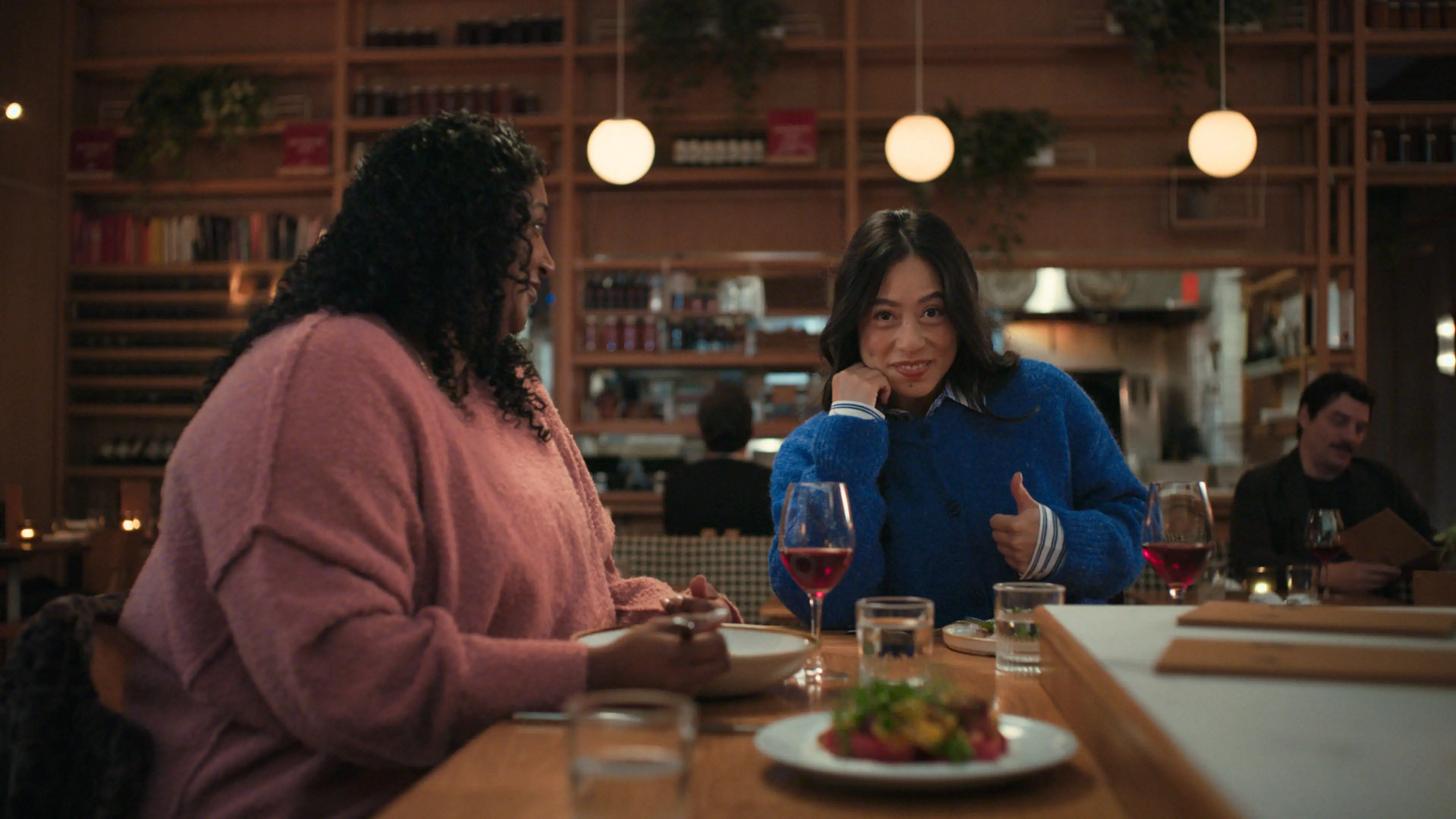Two women sitting at a restaurant table with wine glasses and plates, one woman in a pink sweater and the other in a blue sweater giving a thumbs up.