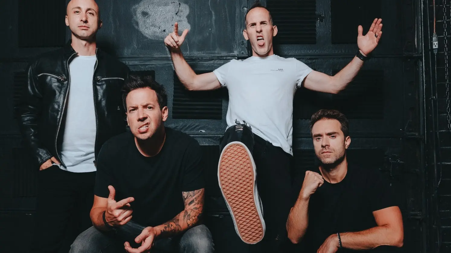 Four people posing against a dark wall, one person in white shirt with foot forward and arms raised, others in black shirts with serious expressions.