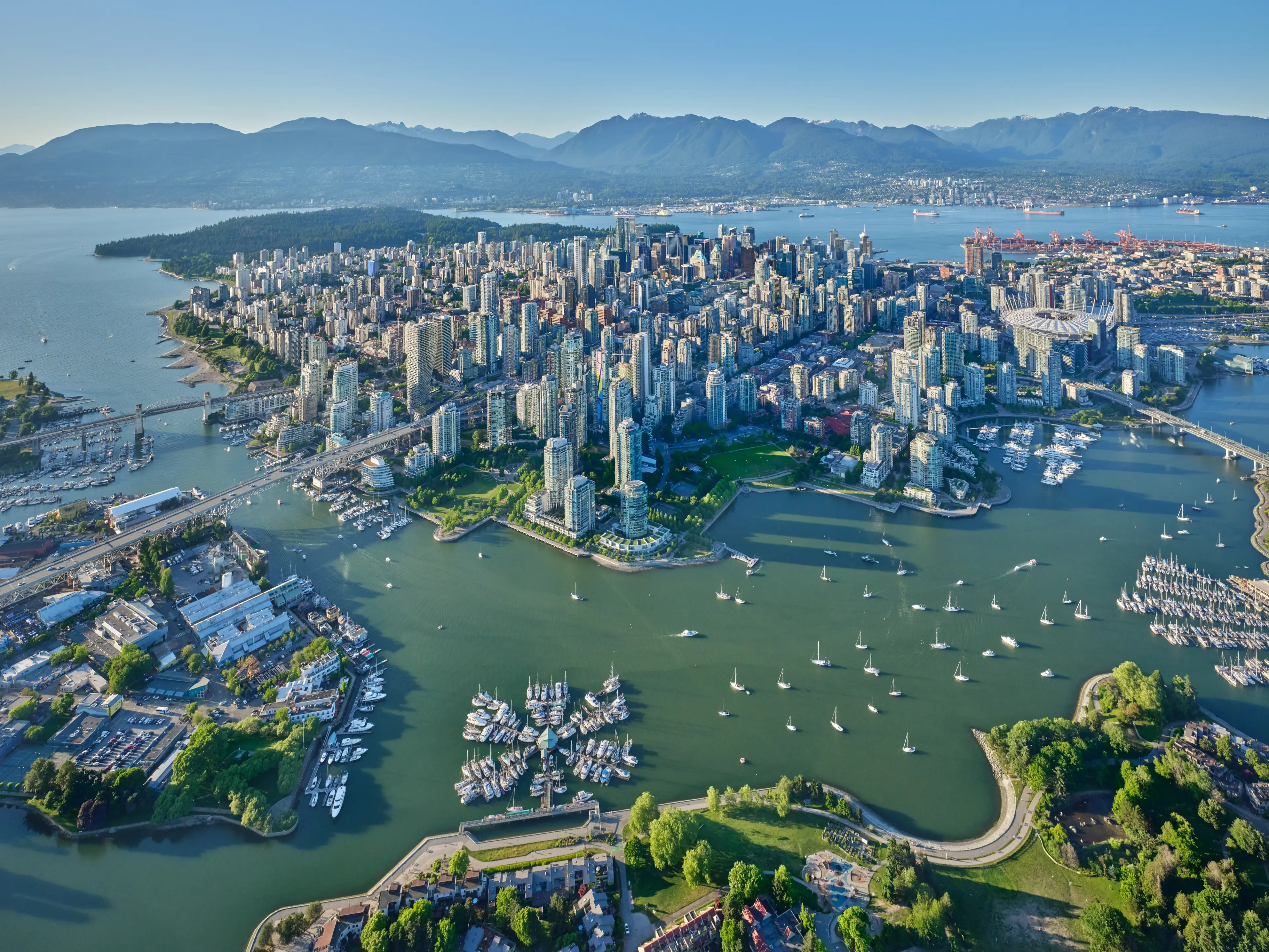 Aerial view of a coastal city with numerous high-rise buildings, surrounded by water with boats and green parks, mountains in the background.