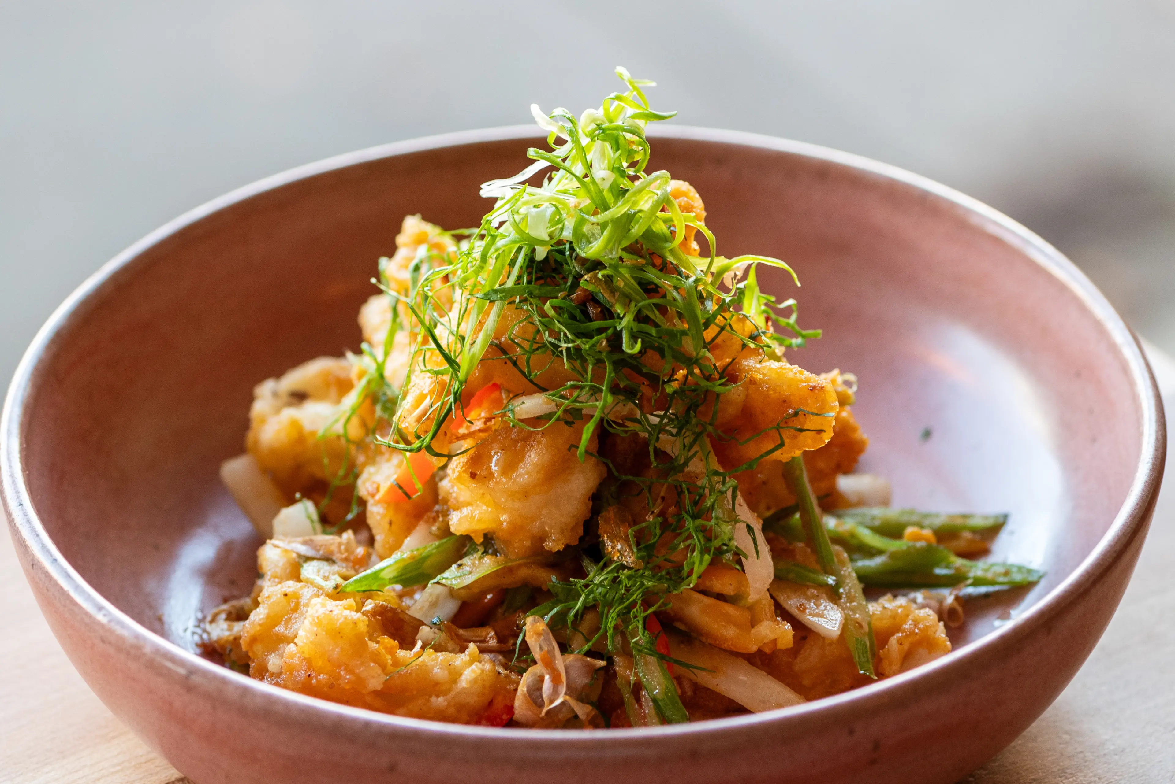 Bowl of fried battered seafood or chicken pieces garnished with green herbs and mixed with sliced vegetables.