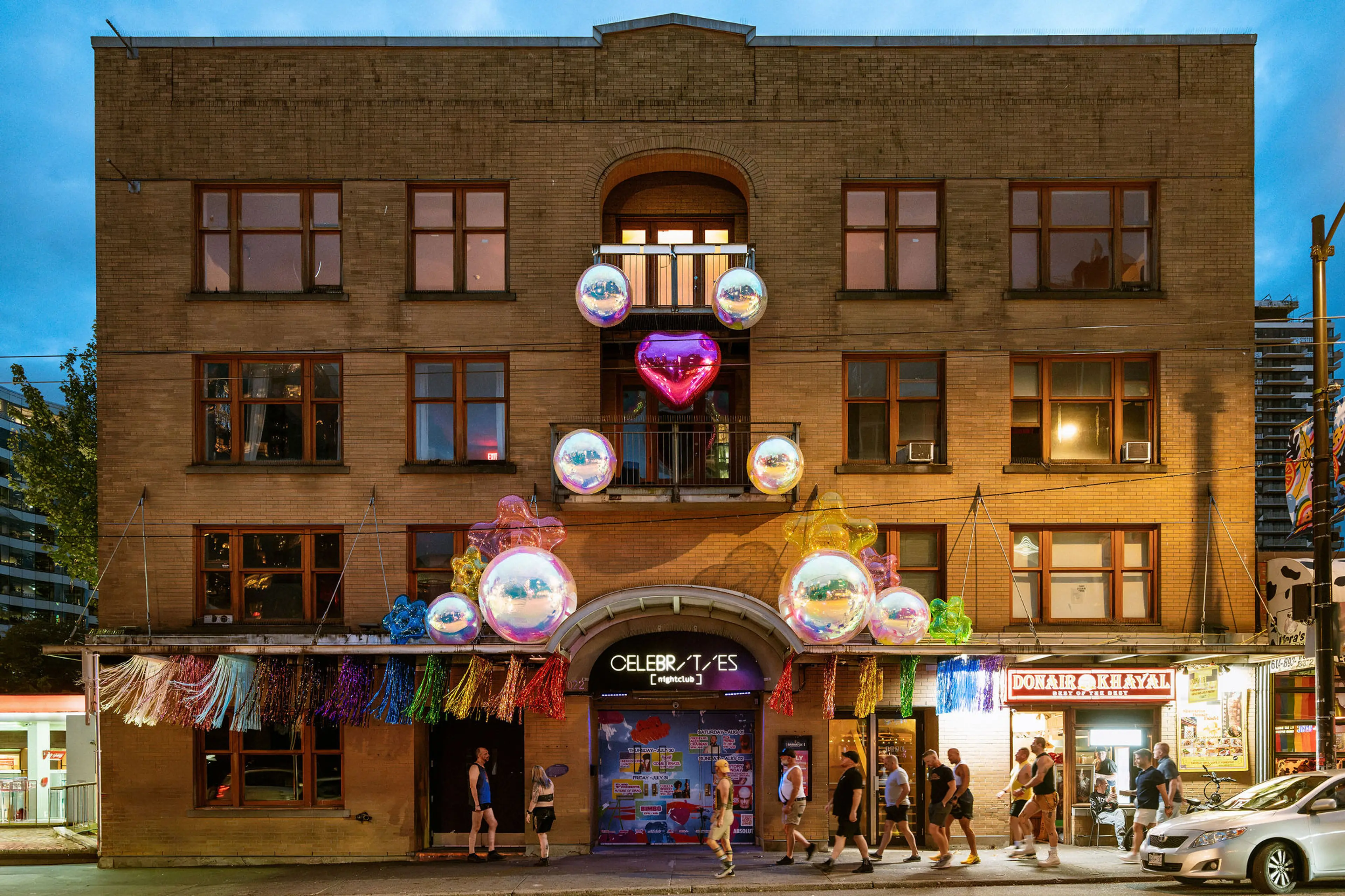 Brick building decorated with colorful balloons and tassels, people walking on the sidewalk in front at dusk.