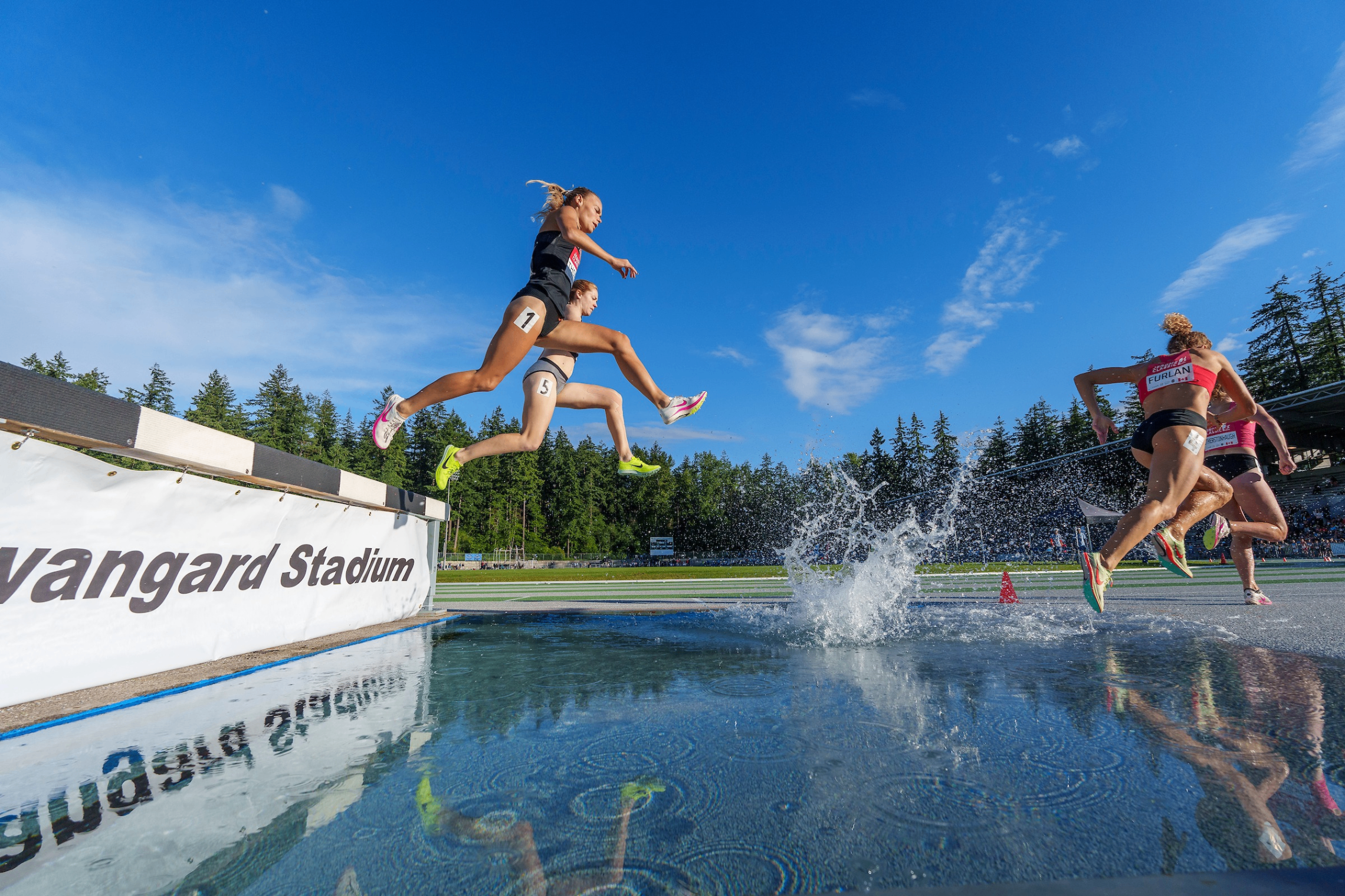 Four athletes jumping over a water obstacle during a steeplechase race at Vanguard Stadium under a clear blue sky.