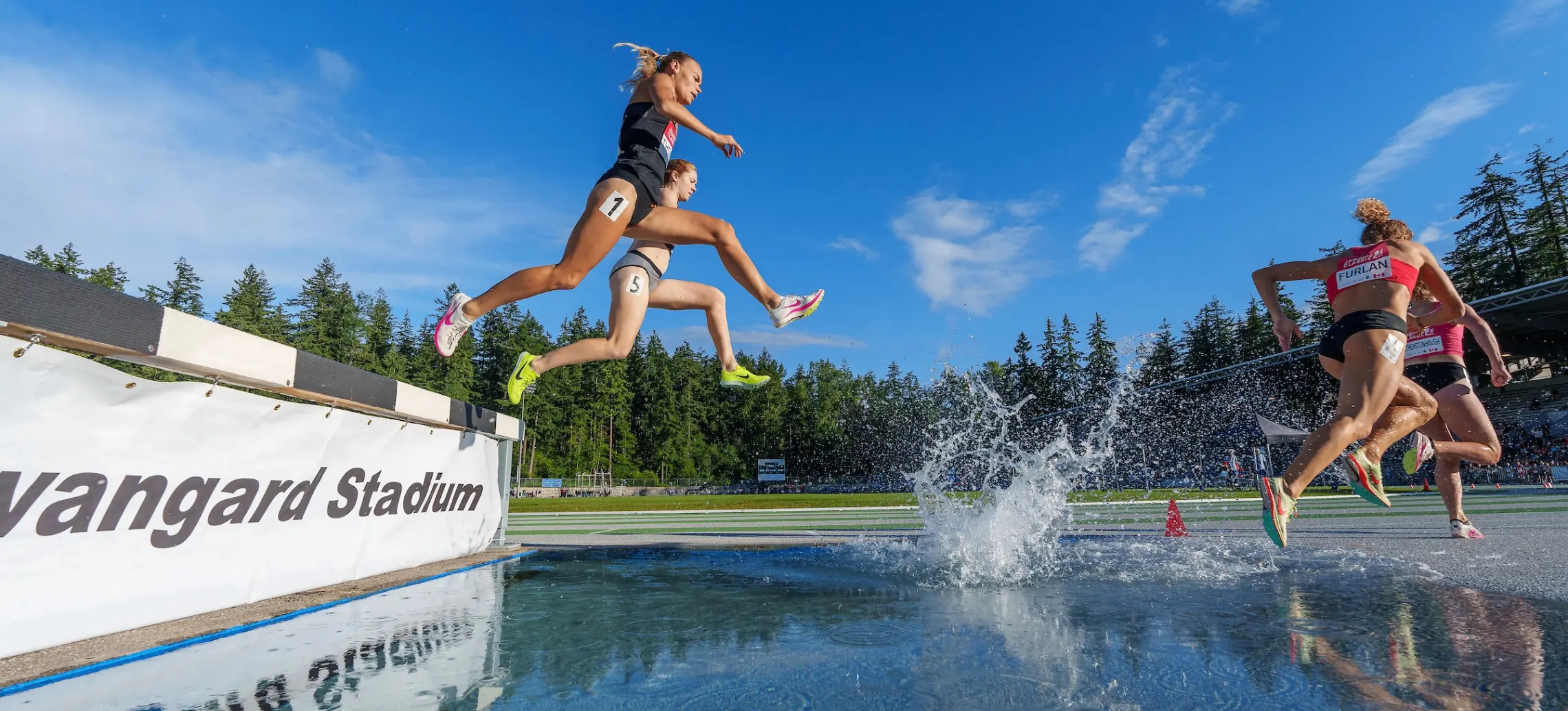 Four athletes jumping over a water obstacle during a steeplechase race at Vanguard Stadium under a clear blue sky.