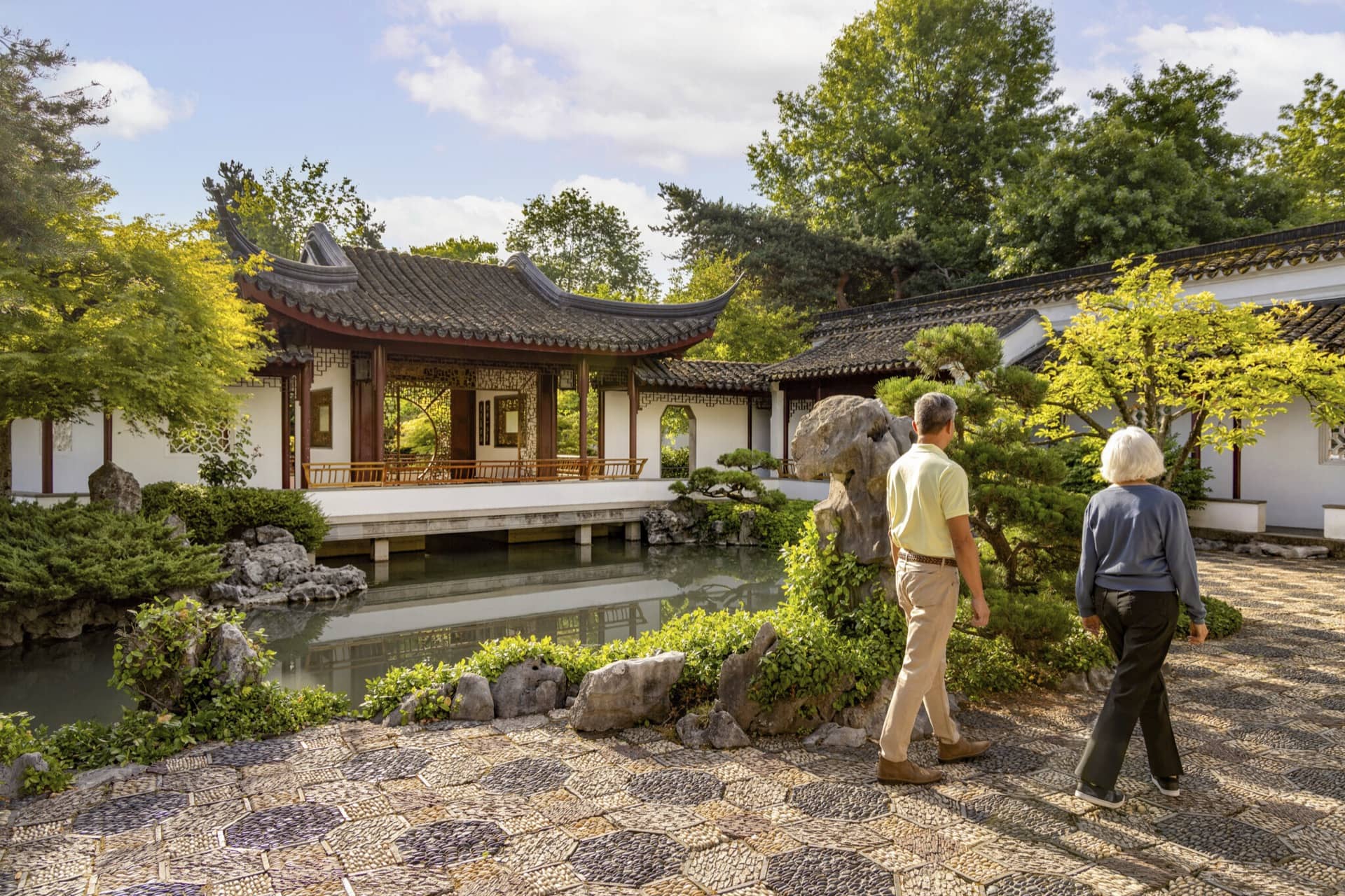Two people walking in a traditional Chinese garden with a pond, rocks, trees, and a pavilion with curved roof.