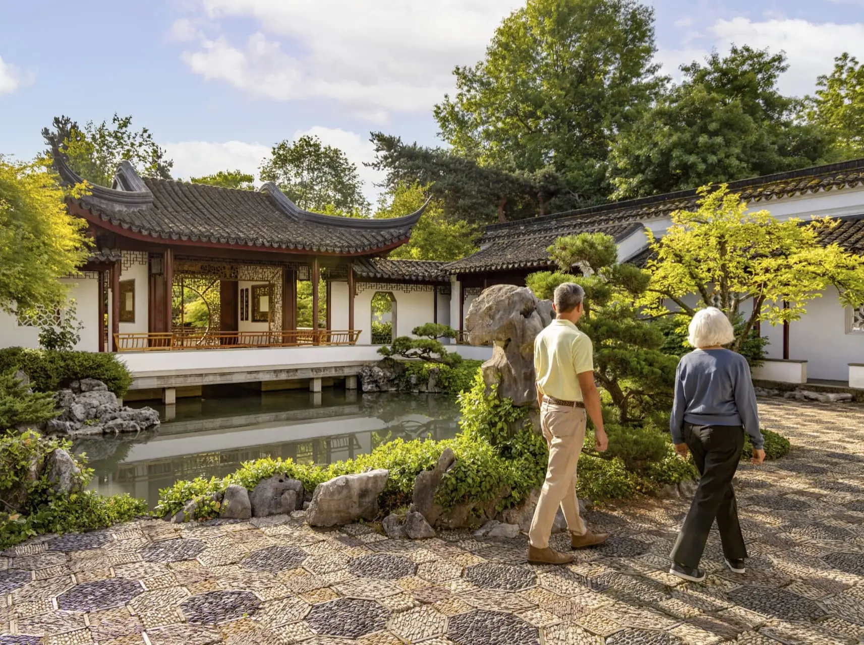 Two people walking in a traditional Chinese garden with a pond, rocks, trees, and a pavilion with curved roof.