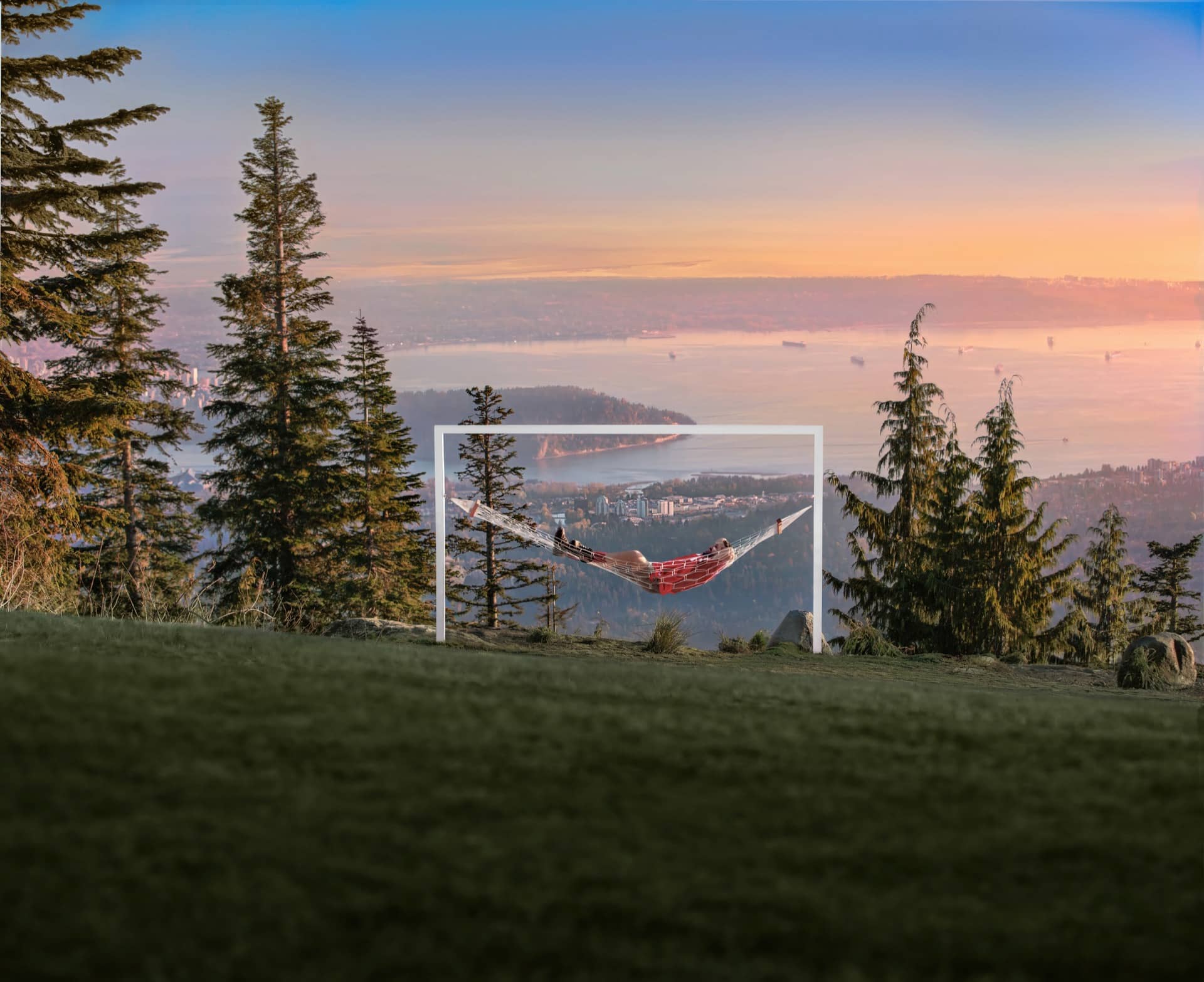 Person relaxing in a hammock on a hilltop with trees and a city and water view at sunset.