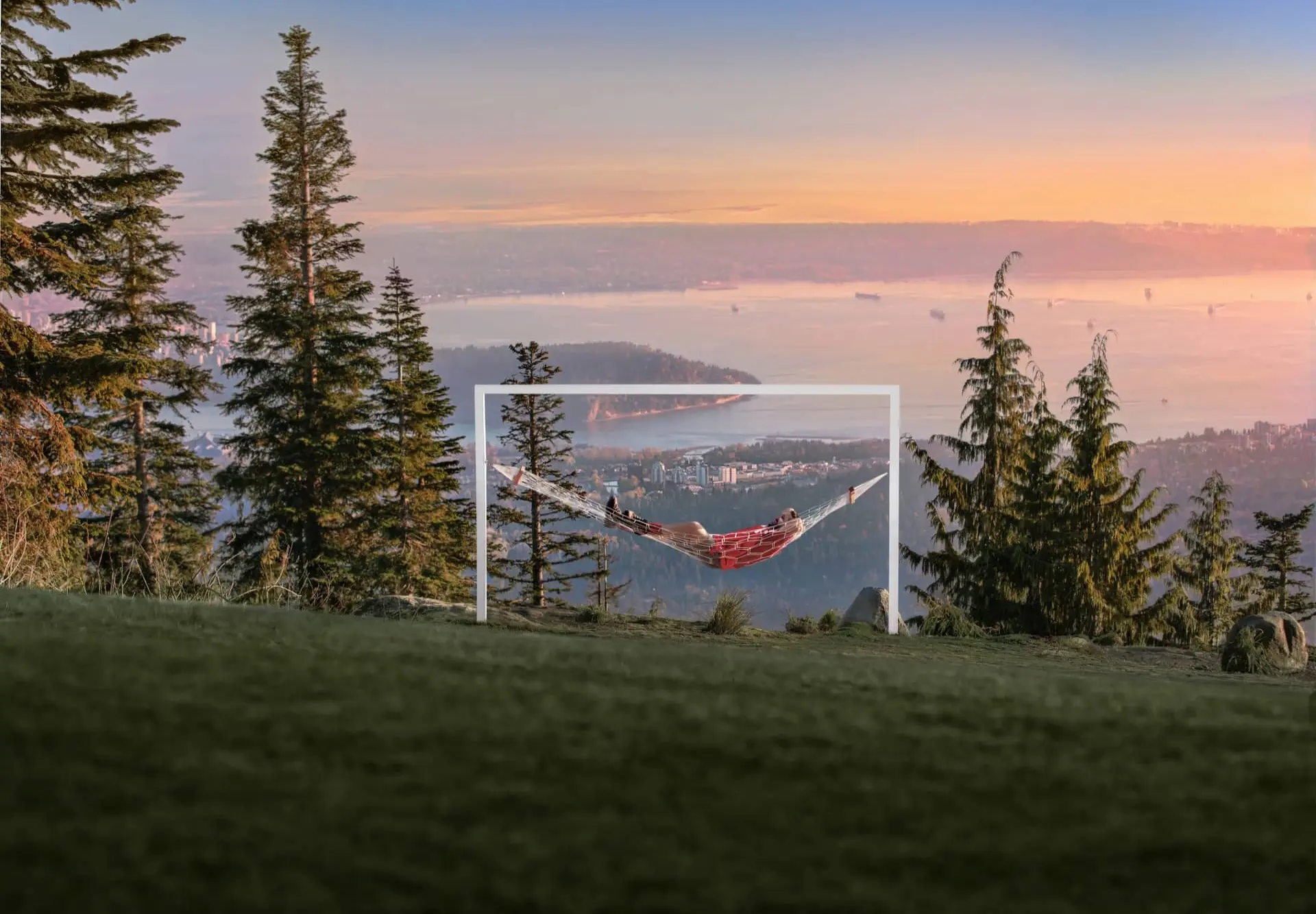 Person relaxing in a hammock on a hilltop with trees and a city and water view at sunset.