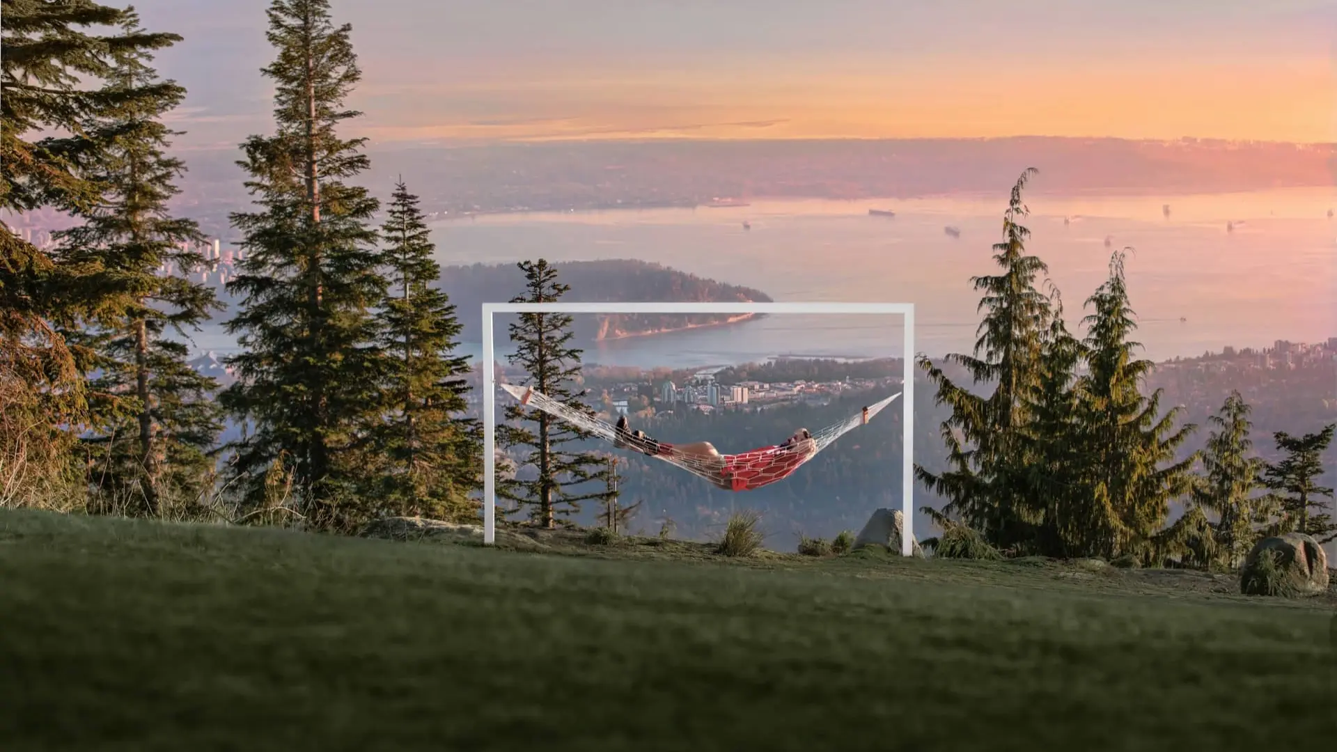 Person lying in a red hammock suspended within a white frame on a grassy hill with trees and a city and water view at sunset.