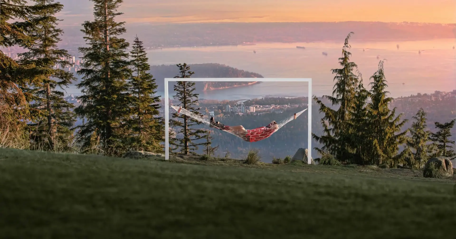 Person lying in a red hammock suspended within a white frame on a grassy hill with trees and a city and water view at sunset.